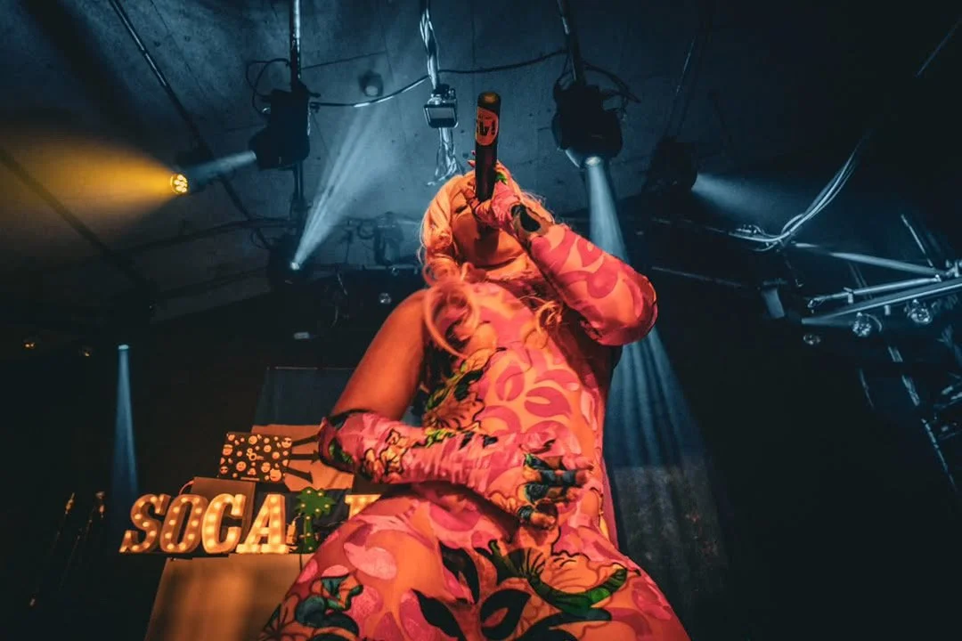 Jadel in colorful, patterned clothing singing into a microphone on stage with stage lights and equipment overhead.