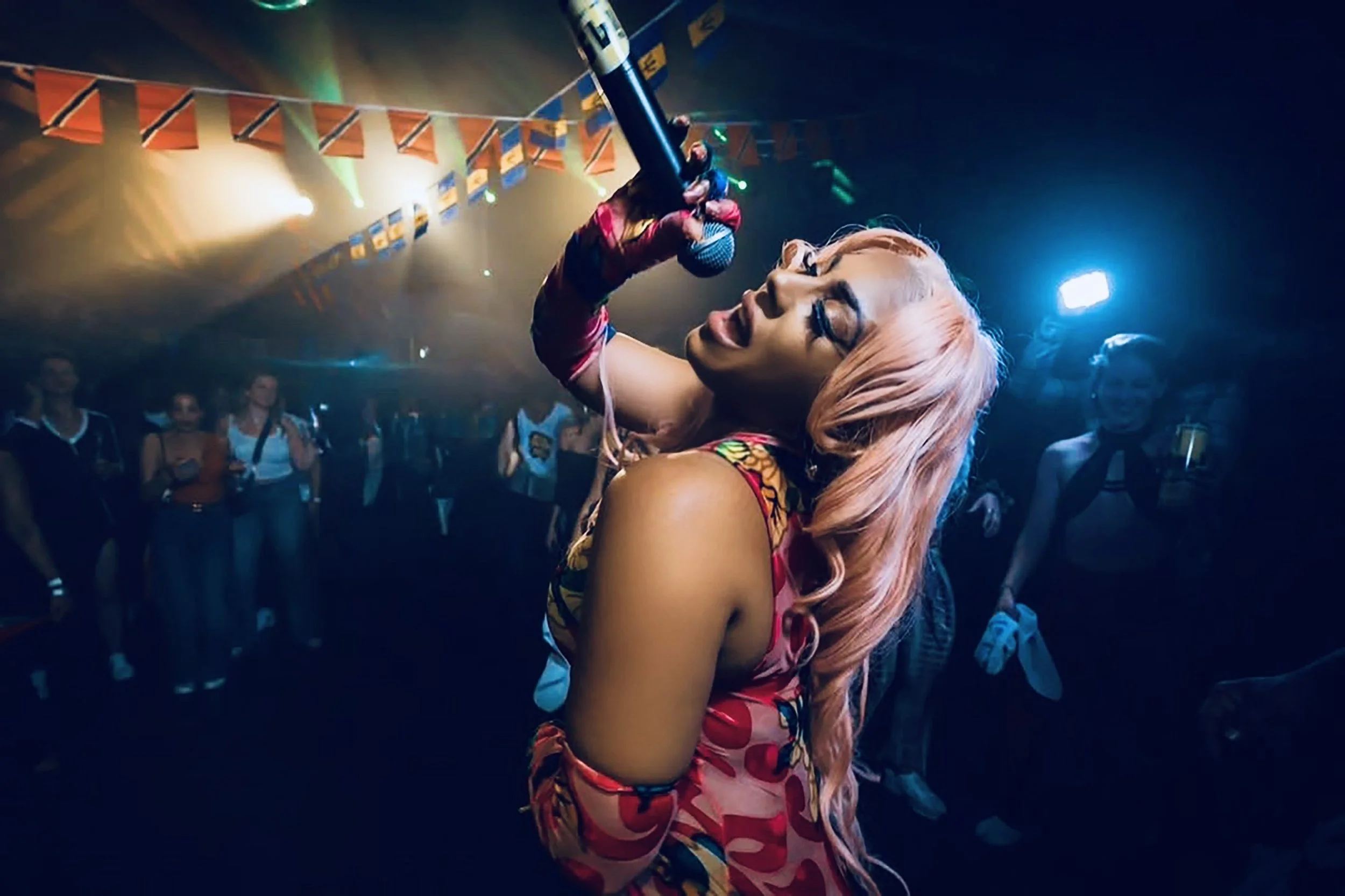 Jadel  with pink hair, glasses, and colorful clothing holding a microphone and singing on stage at a lively indoor concert with fog and colorful lights.