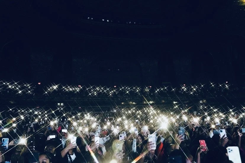 Jadel in front a Crowd of people at a concert or event, holding up phones with flashlights on, creating a starburst effect in the dark.