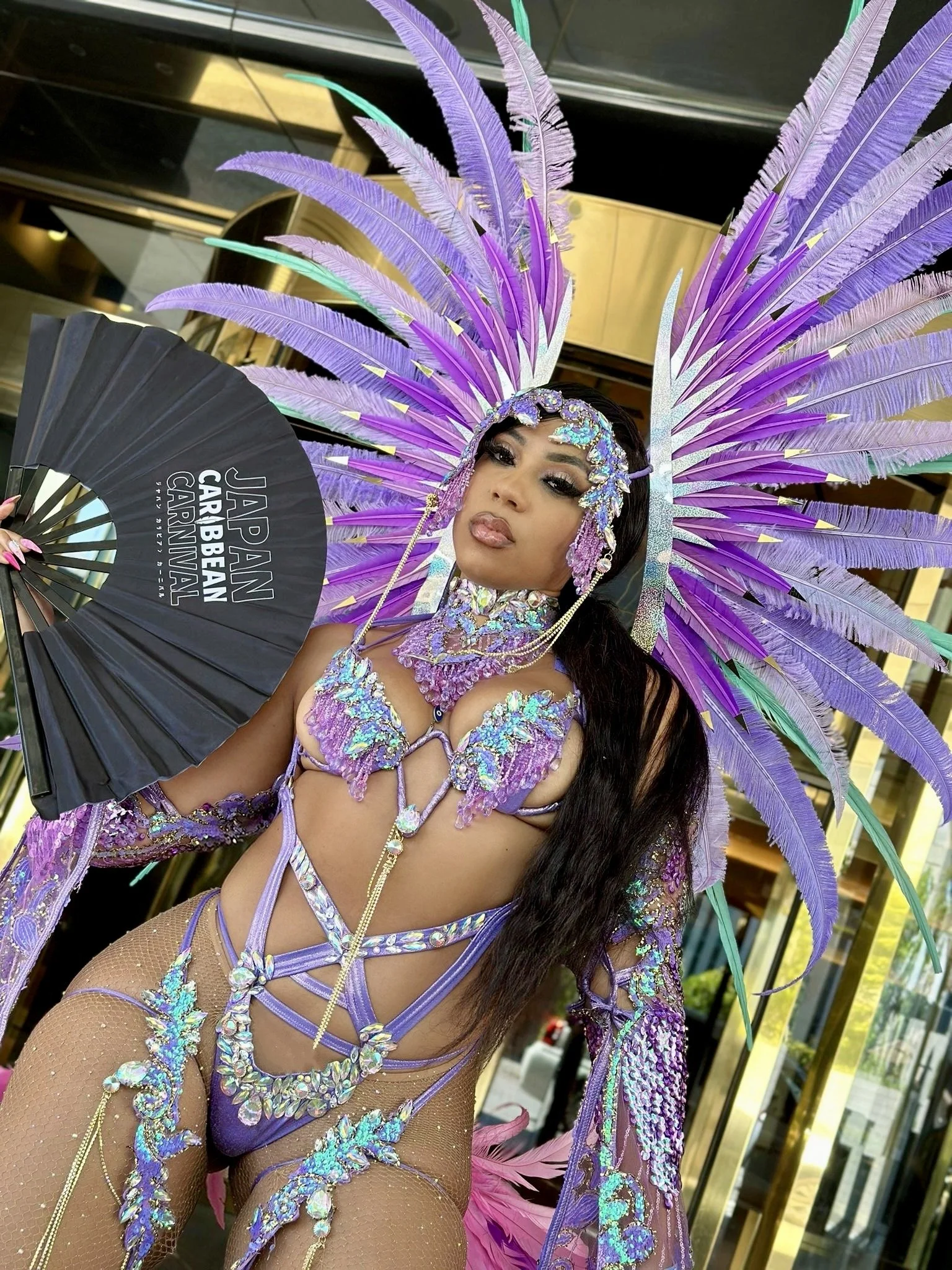 A woman dressed in a colorful, elaborate carnival costume with purple and pink feathers, rhinestones, and sequins, holding a black fan with white text reading 'JAPAN CARNIVAL' in front of a building with glass windows.