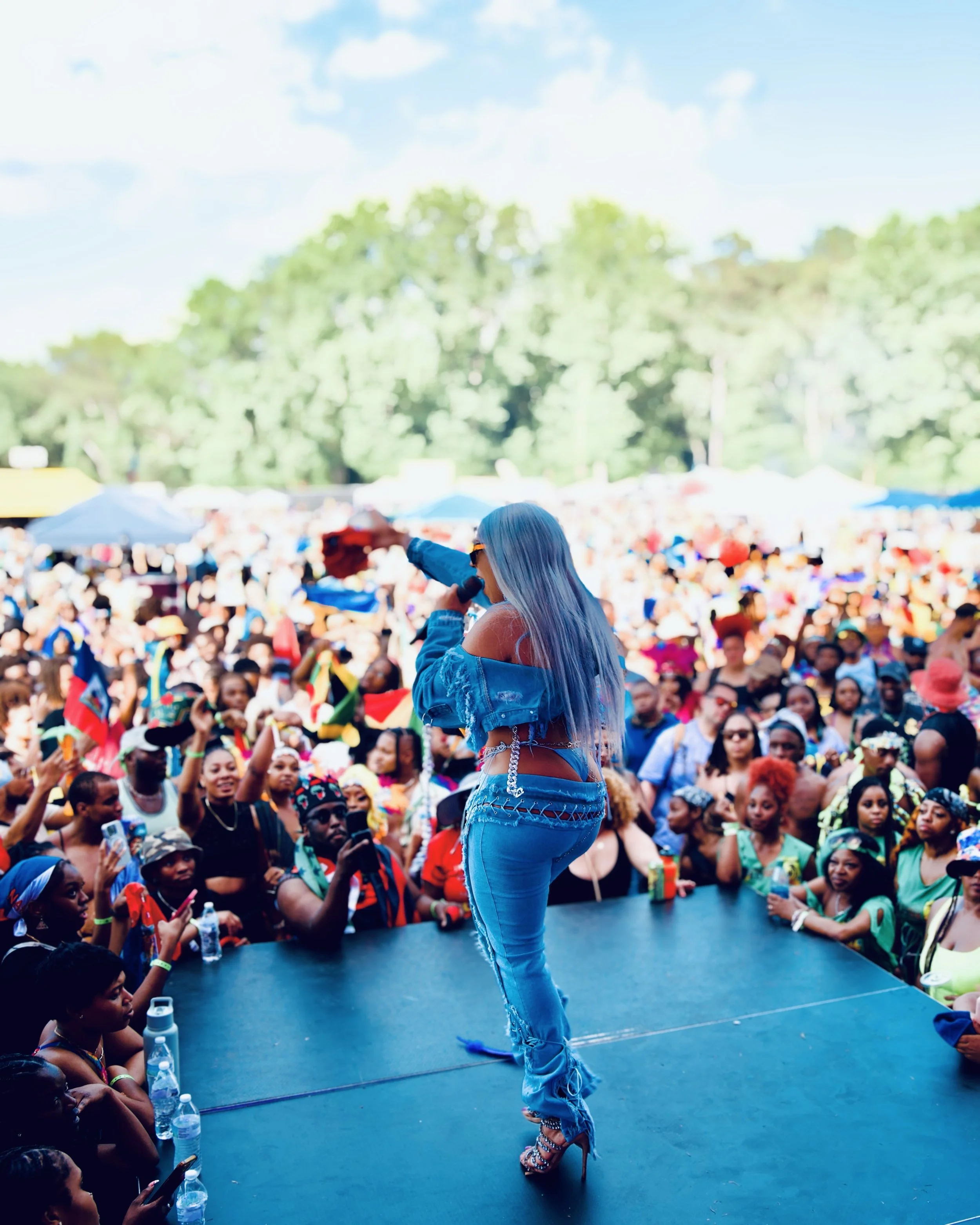 Jadel with long gray hair and sunglasses stands on stage facing an audience at an outdoor event. The audience is seated and engaged, with many holding cameras or phones. Atlanta Carnival 2024