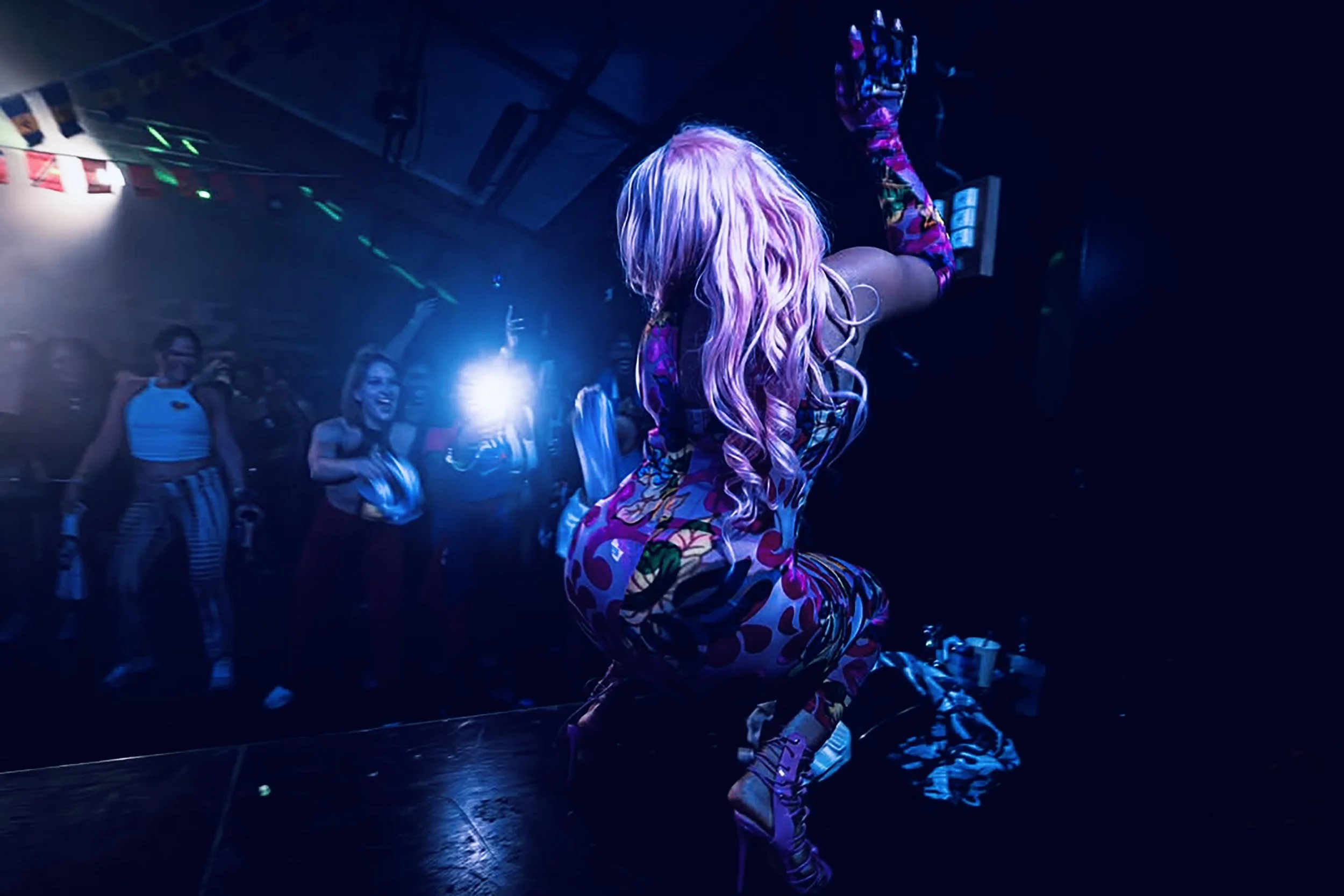 Jadel with long, wavy pink hair wearing a colorful, patterned outfit is dancing or posing on a dark stage illuminated by vibrant, colorful lights. Audience members are visible in the background, some taking photos or videos.
