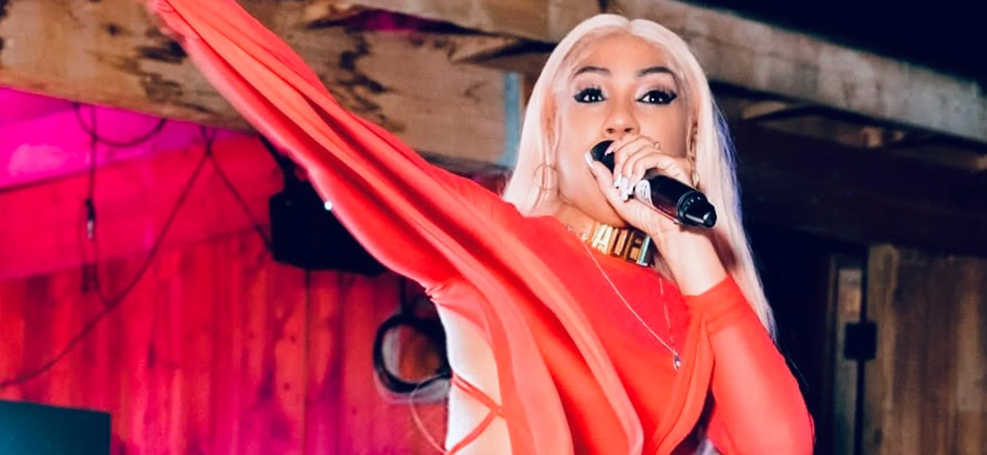 Jadel  with platinum blonde hair singing into a microphone with her arm raised, dressed in a red outfit, in a dimly lit venue with red curtains in the background.