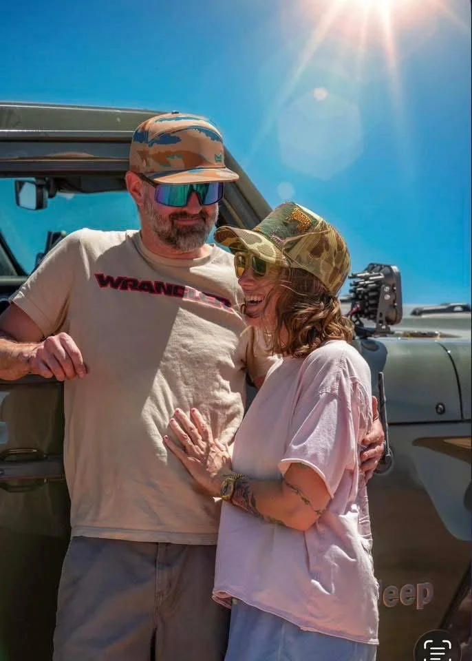 A man and woman smiling and enjoying each other's company outdoors during sunny weather. They are wearing camouflage hats and sunglasses, with a rugged vehicle, possibly a Jeep, in the background.