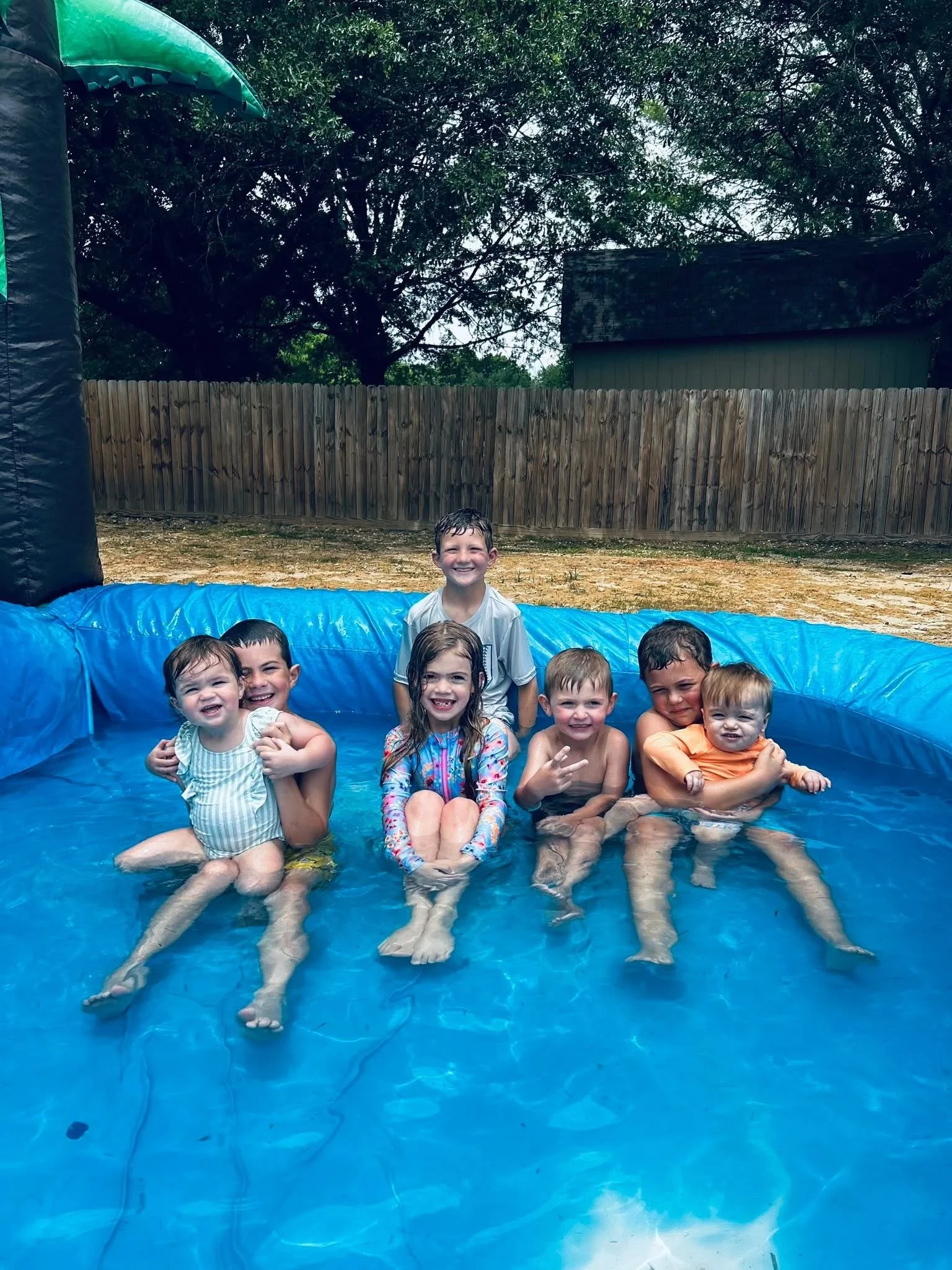 Group of children sitting in a blue inflatable pool outdoors, smiling, with trees and a wooden fence in the background.
