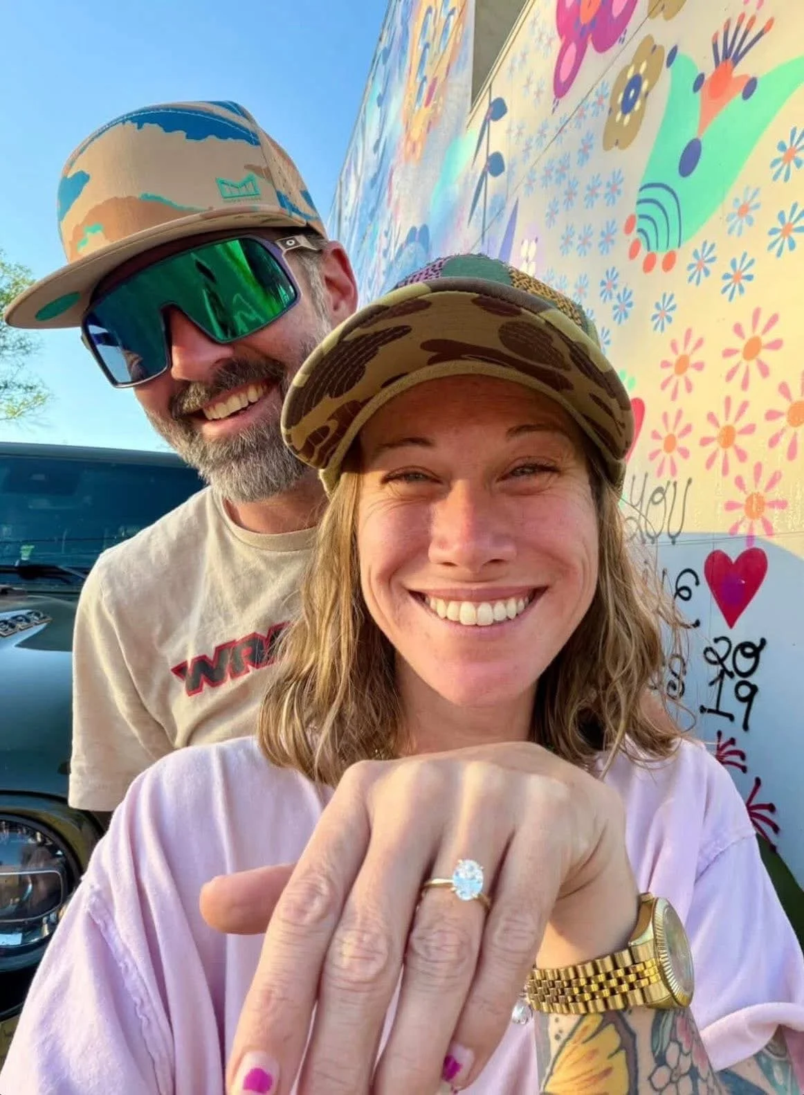 A smiling woman showing off her engagement ring, with a man smiling behind her. They are outdoors, with a colorful mural visible in the background.