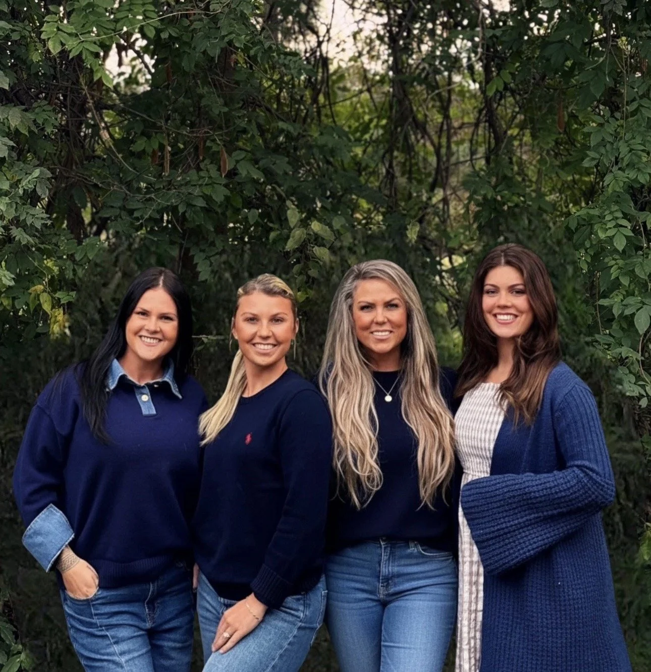 Four women standing outdoors in front of green leafy trees, smiling, dressed in casual blue and black outfits.