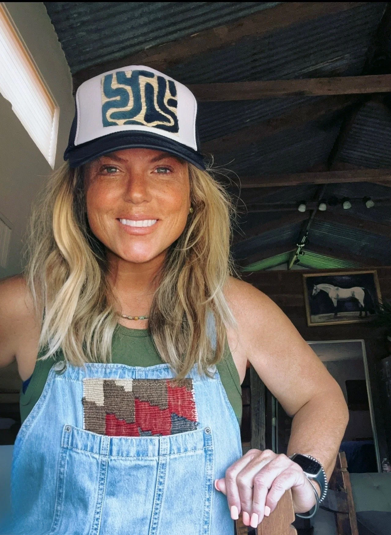 A woman with blonde wavy hair wearing a white and black trucker hat, a green tank top, and denim overalls with a patchwork design, smiling inside a rustic room with wooden ceiling beams and a framed horse picture in the background.