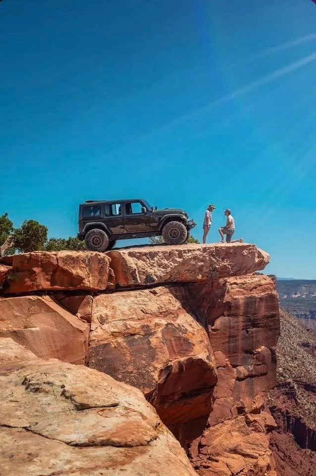 A black SUV parked on the edge of a rocky cliff with two people standing nearby, overlooking a canyon under a clear blue sky.