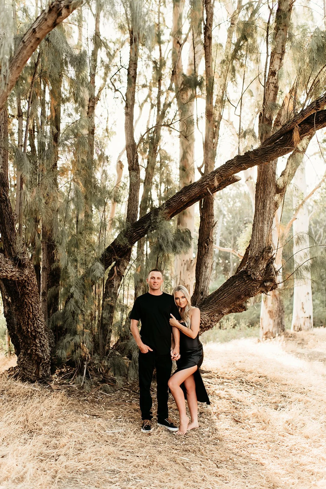 A man and woman standing together outdoors in a wooded area during daytime, with the woman wearing a black dress and the man in a black t-shirt and pants.