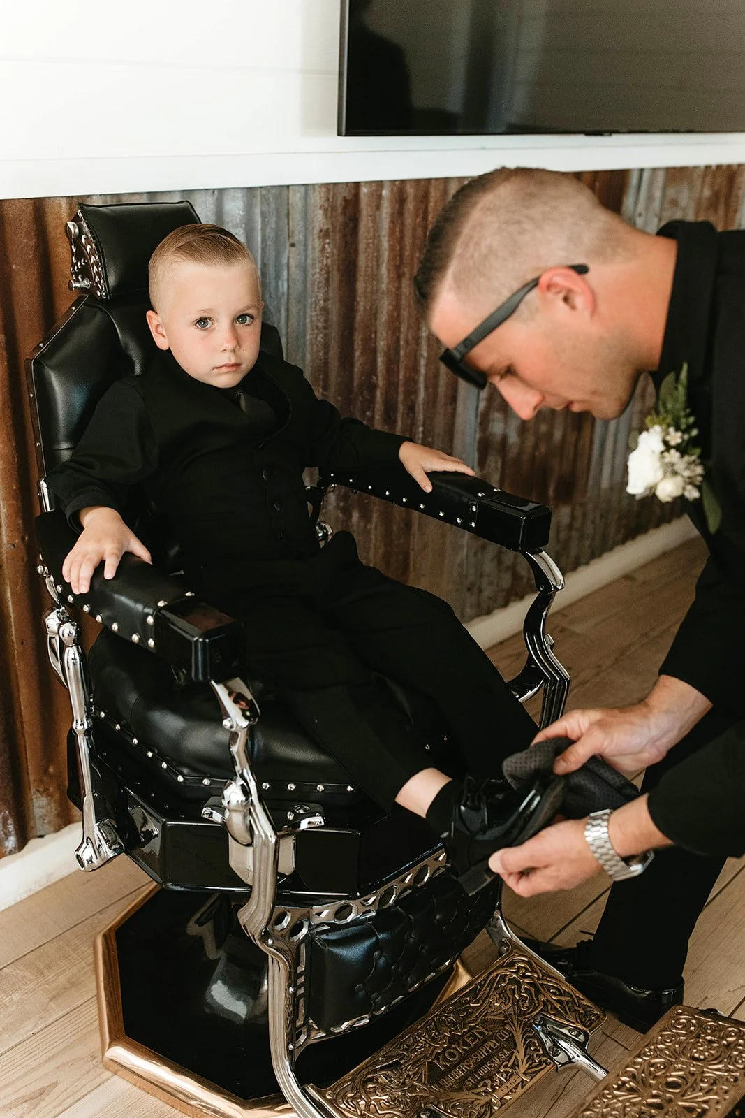 Young boy dressed in formal black sitting in a black chair, with a man kneeling beside him adjusting his shoe.