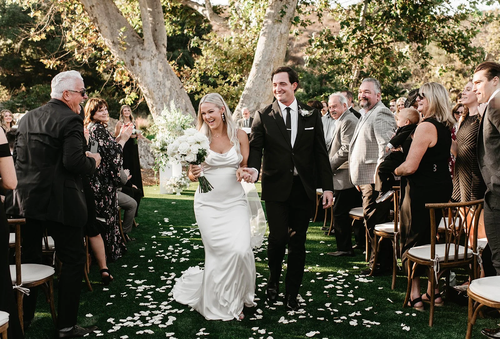 A bride in a white wedding gown and a groom in a black suit walk hand-in-hand down an outdoor aisle lined with seated guests, some clapping and smiling, on a sunny day with trees in the background. White flower petals scatter on the grass.