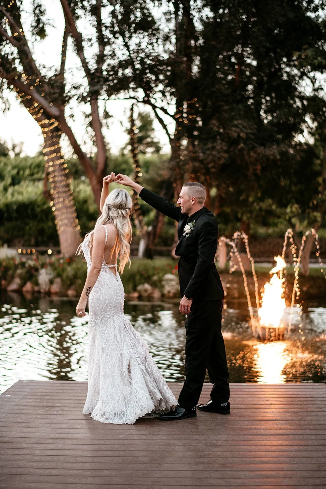 A bride and groom dancing on a wooden dock by a pond during sunset. The bride wears a white lace wedding dress, and the groom wears a black suit. In the background, trees are decorated with string lights and a fountain with water and flames is visibl