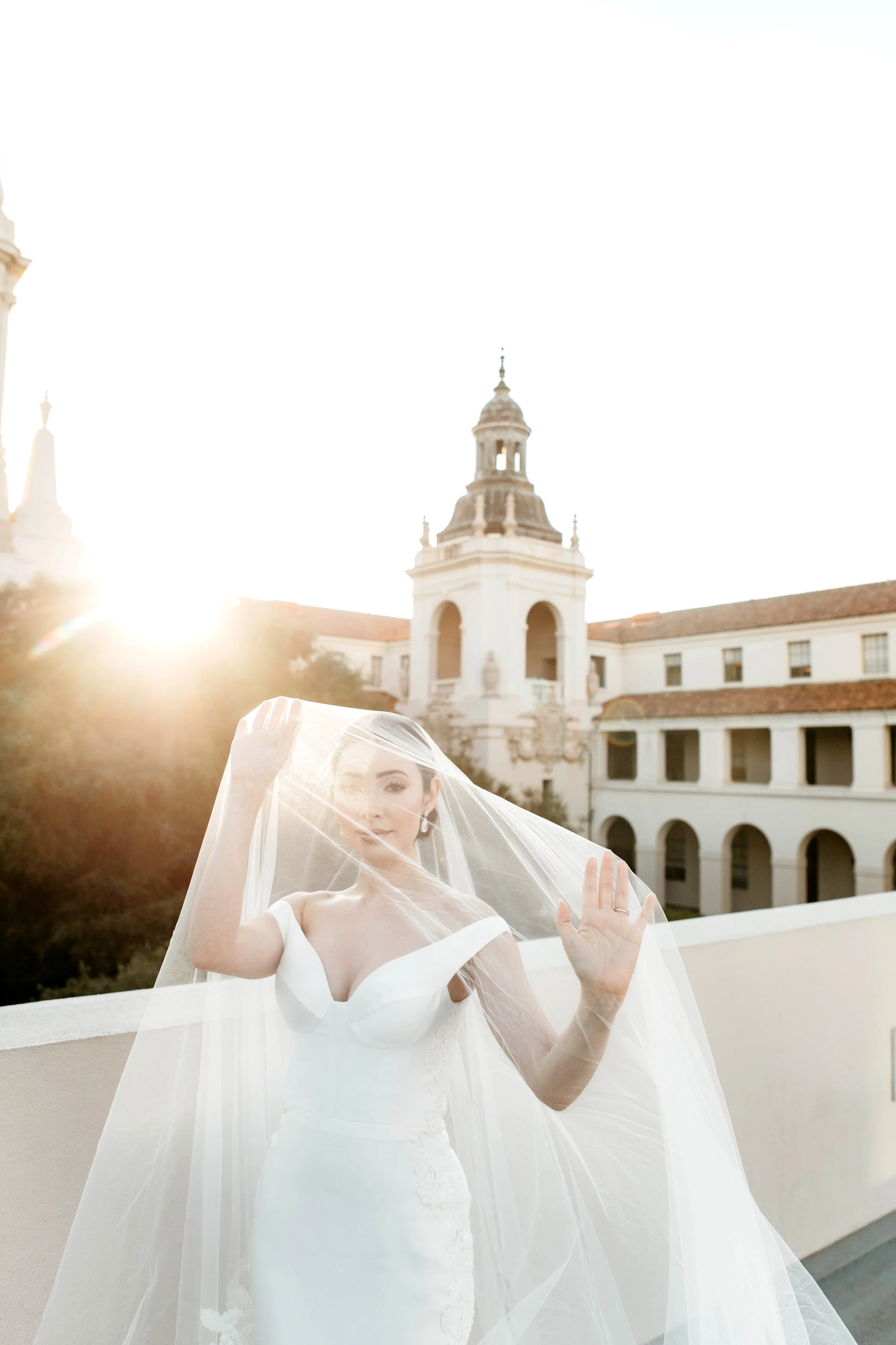 A woman in a white wedding dress and veil standing outdoors near a white building with a bell tower, backlit by the setting sun.