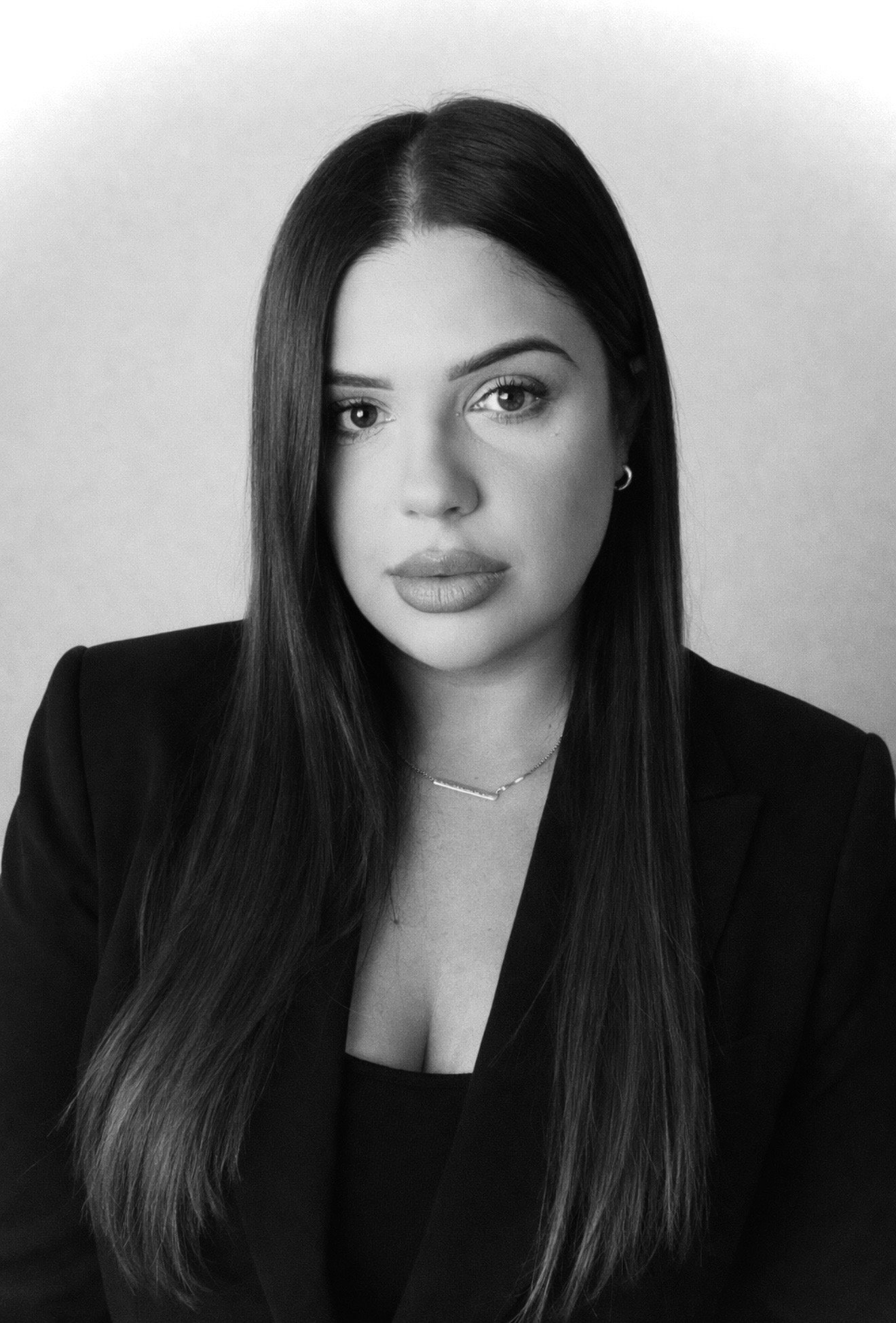 A black and white portrait of a woman with long, straight hair, wearing a black blazer and a necklace, looking directly at the camera.