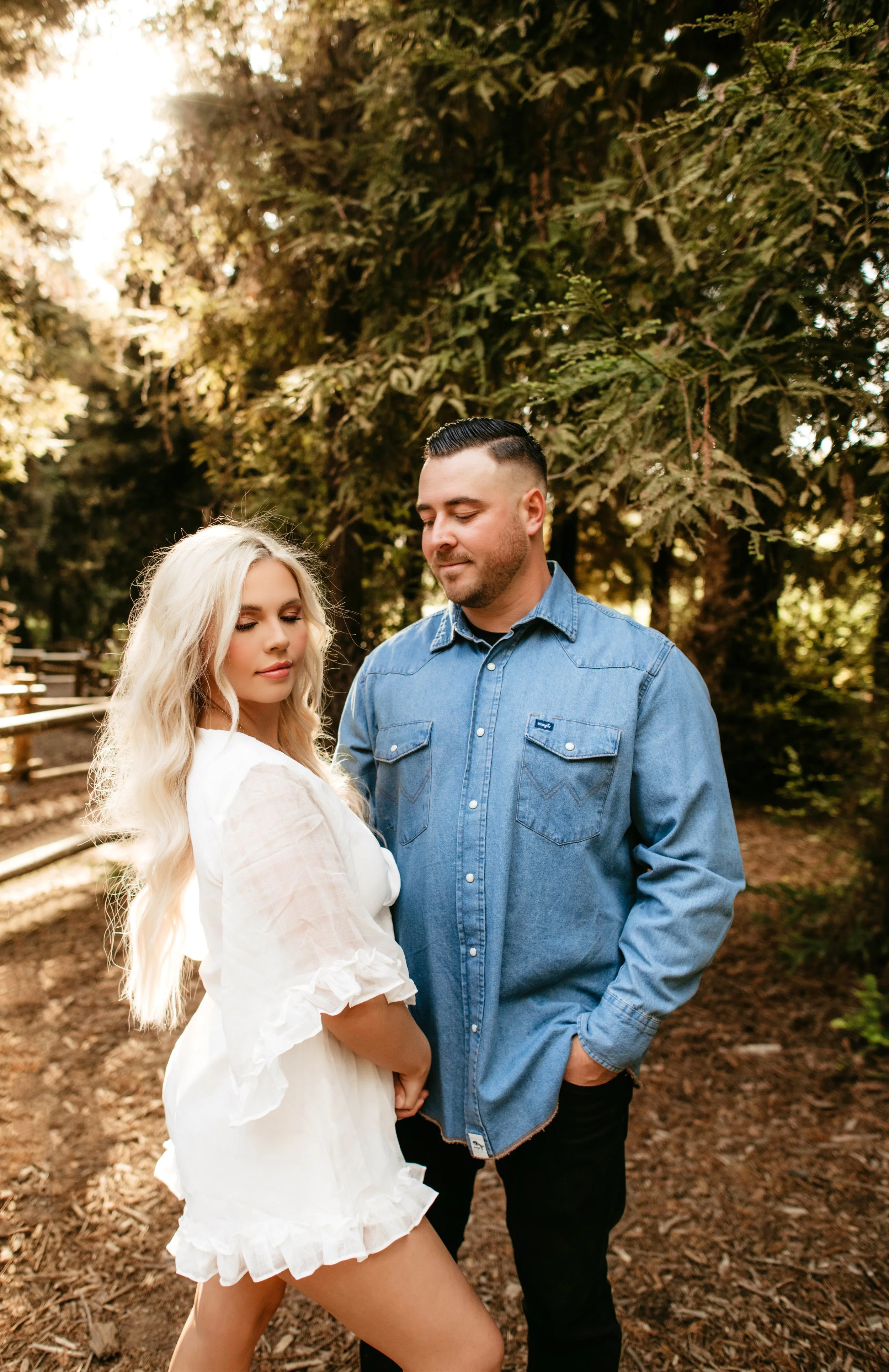 A couple standing outdoors beneath trees, the woman with long blonde hair wearing a white dress, and the man with short dark hair wearing a denim shirt, holding hands and looking content.