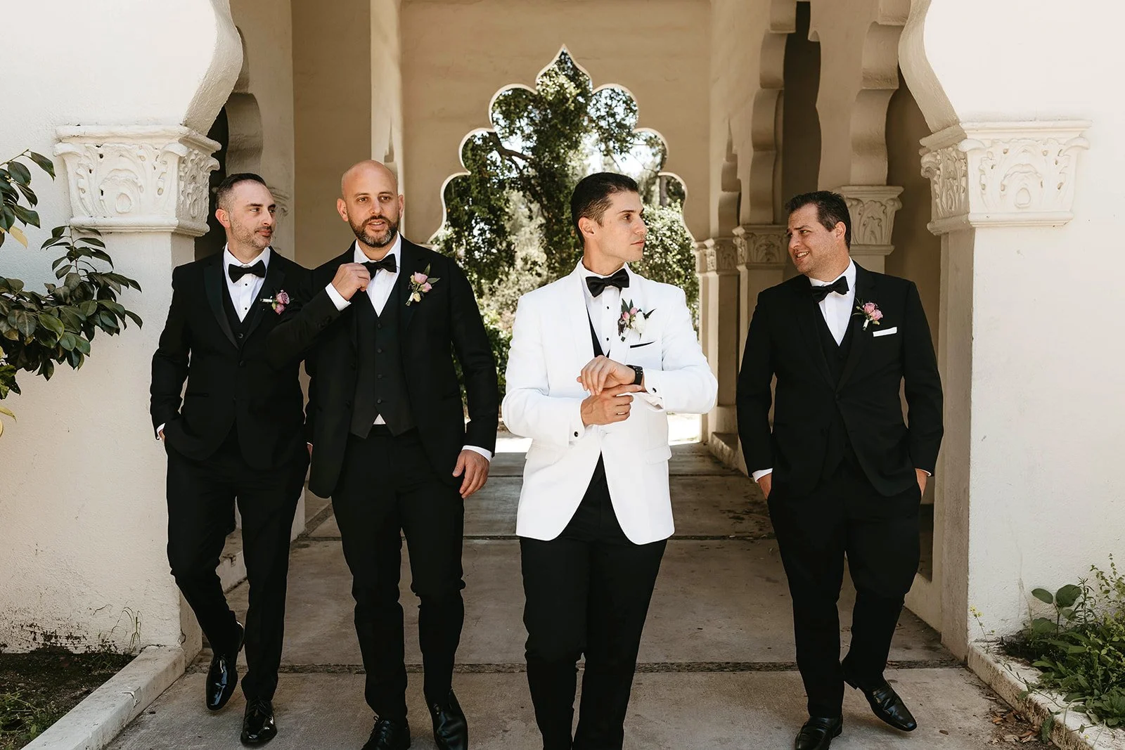 Five men dressed in tuxedos, one in a white tuxedo, walking and posing outdoors near a decorative archway.