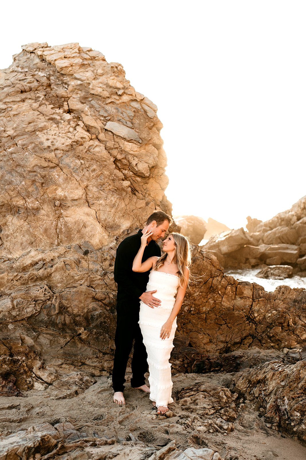 A romantic couple standing close together on a rocky beach at sunset, with large rocks in the background, gazing into each other's eyes.