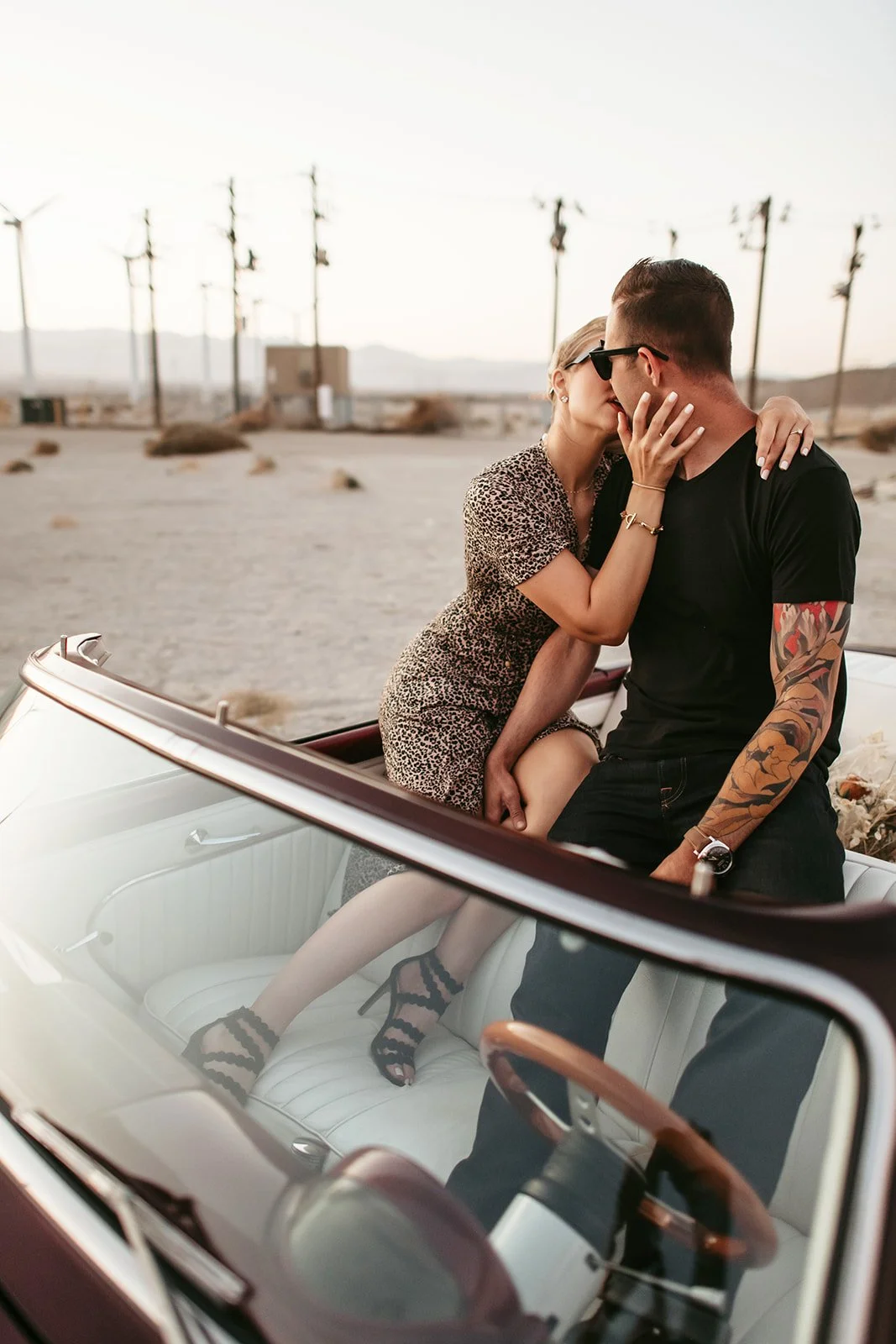 A couple sharing a kiss inside a vintage car in a desert landscape with power lines in the background.
