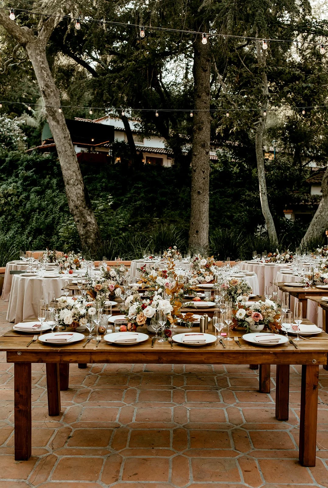 Outdoor banquet table decorated with floral arrangements, set with plates, glasses, and silverware in a garden setting with trees and string lights overhead.