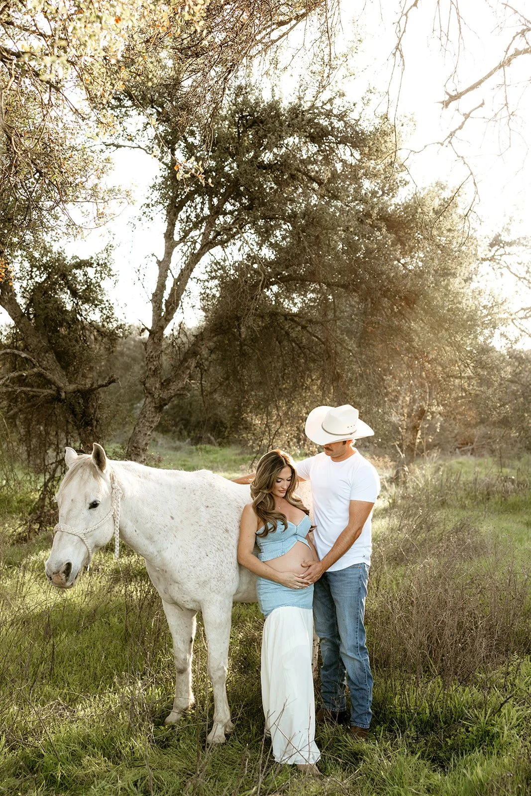 A pregnant woman and a man are standing outdoors with a white horse. The woman is cradling her baby bump with her hands, while the man places one hand on her belly and the other on her back. They are near trees, and sunlight filters through the branc