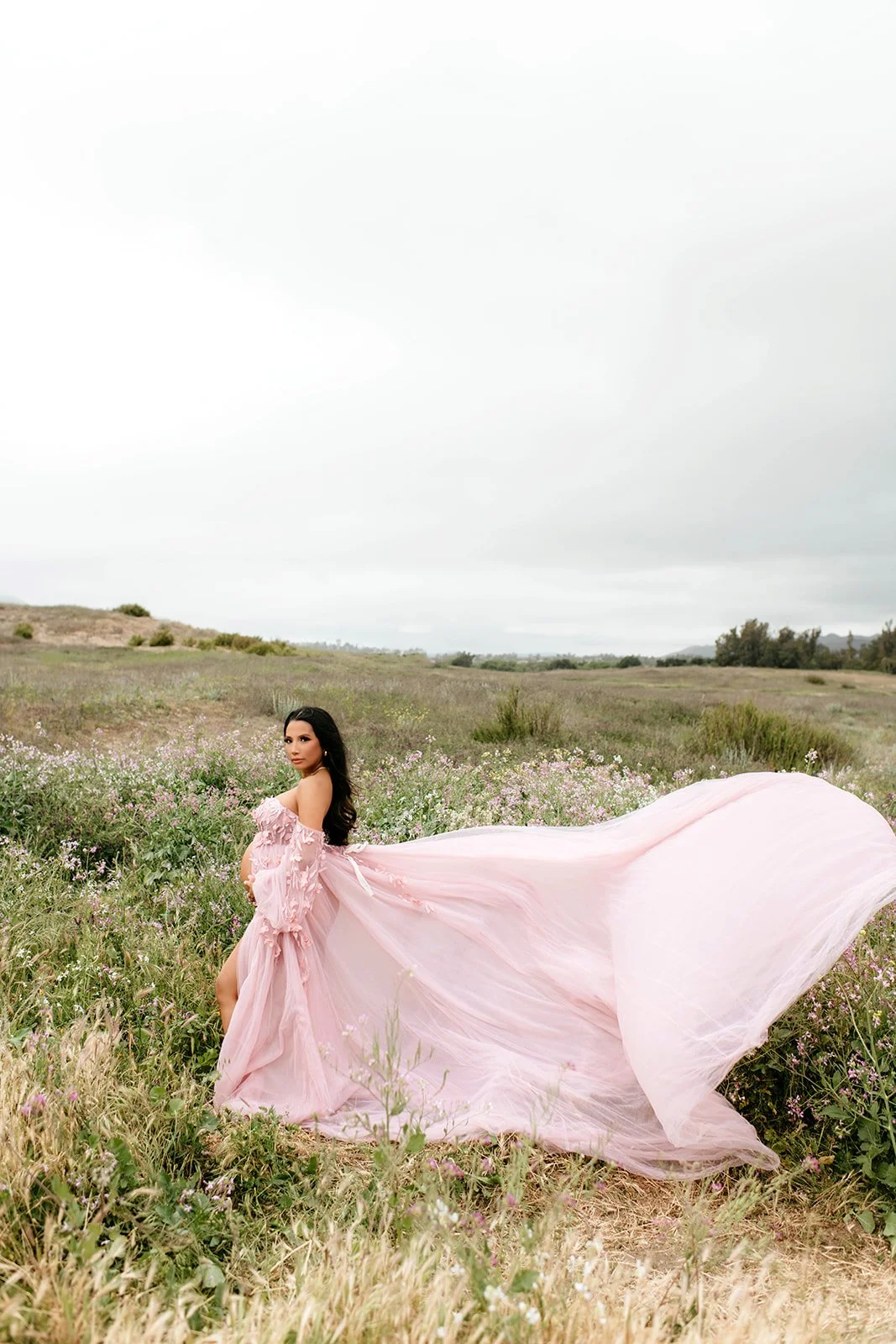 A woman in a flowing pink dress standing in a field of wildflowers under a cloudy sky.