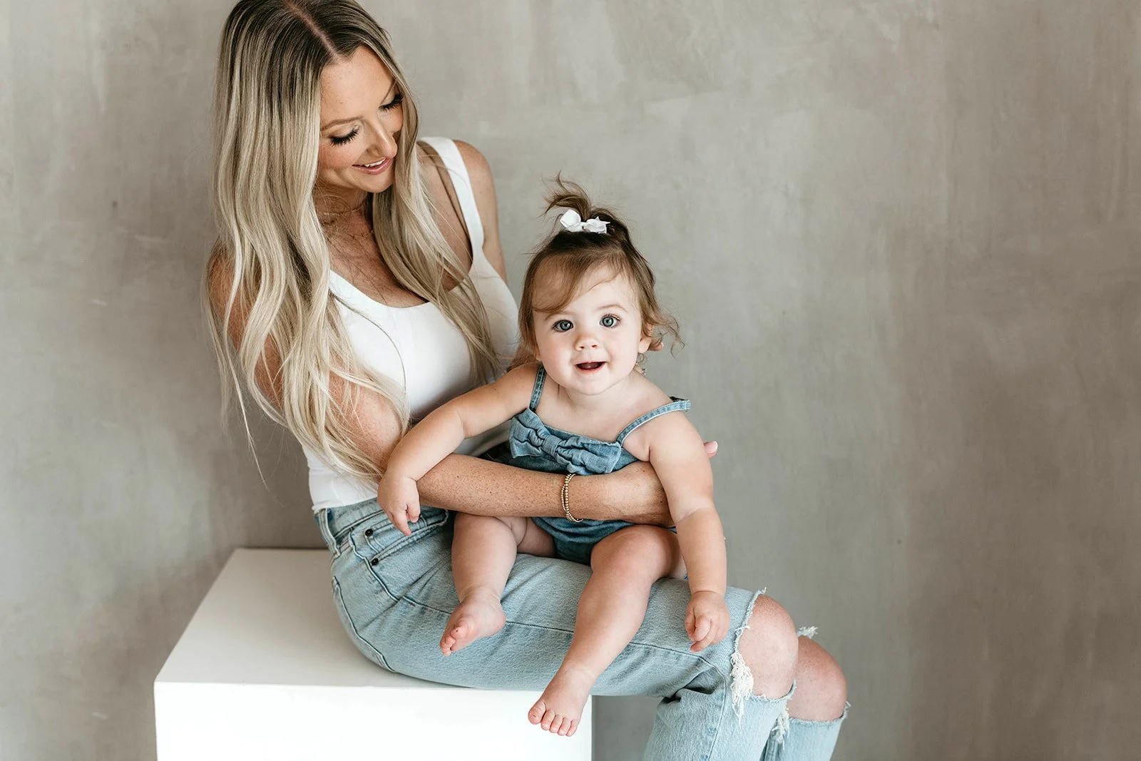 A woman with long blonde hair sitting on a white platform holding a young girl with curly hair and a white bow, both smiling against a plain background.