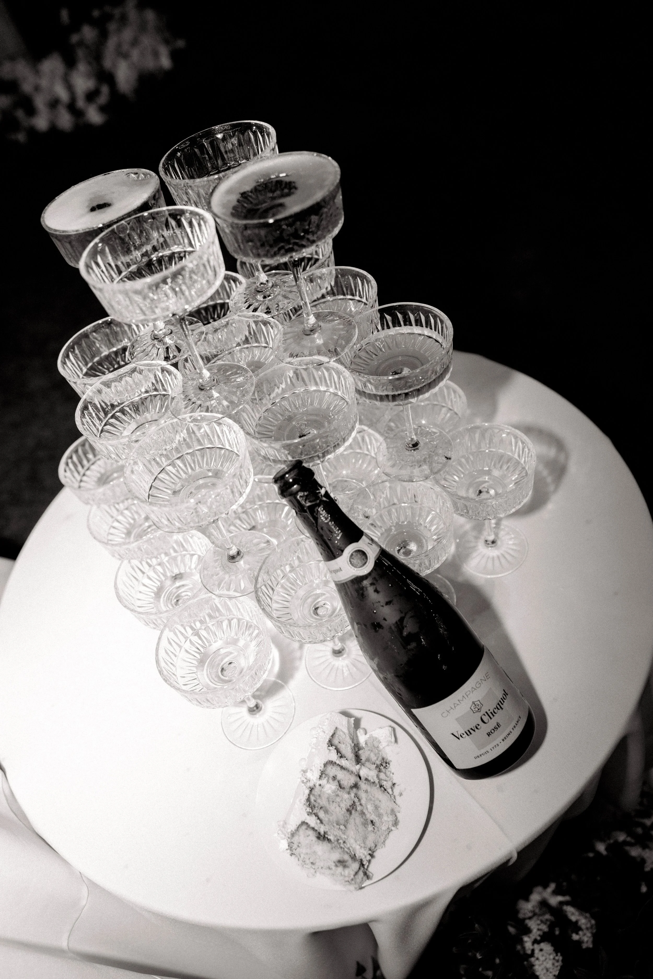 A tiered display of empty crystal champagne glasses on a white table, with a bottle of Veuve Clicquot Rose champagne and a partially eaten slice of cake on the table.