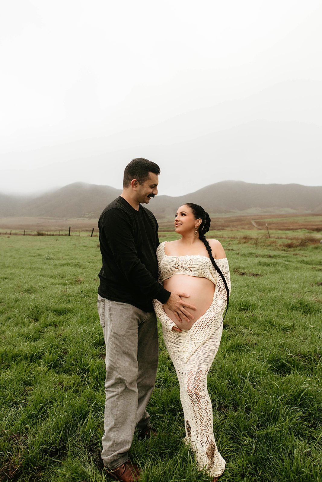 A couple standing on green grass in a field, the pregnant woman is wearing a cream crocheted dress and the man is in a black shirt and khaki pants; they are smiling and tenderly touching her baby bump, with hills in the background and a cloudy sky.