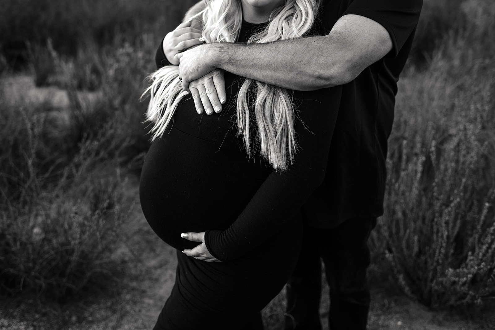 A pregnant woman with long hair being embraced from behind by a man, both wearing black attire, standing outdoors in a natural setting with tall grass.