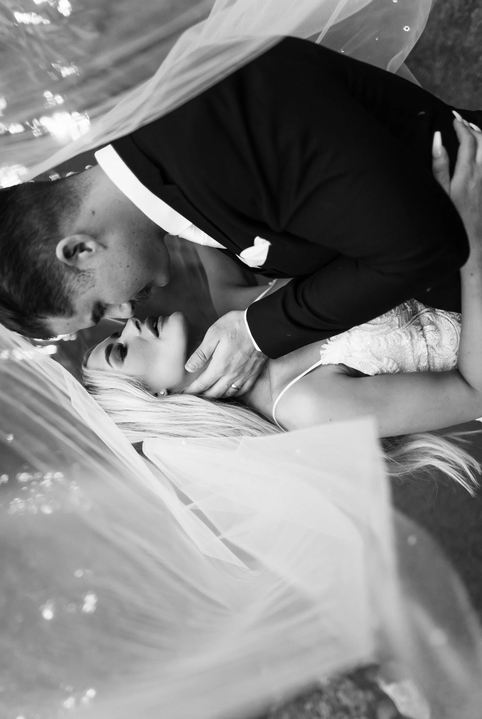 A man and a woman sharing a kiss, with the man dressed in a tuxedo and the woman in a wedding dress, seen through a plastic or glass barrier.