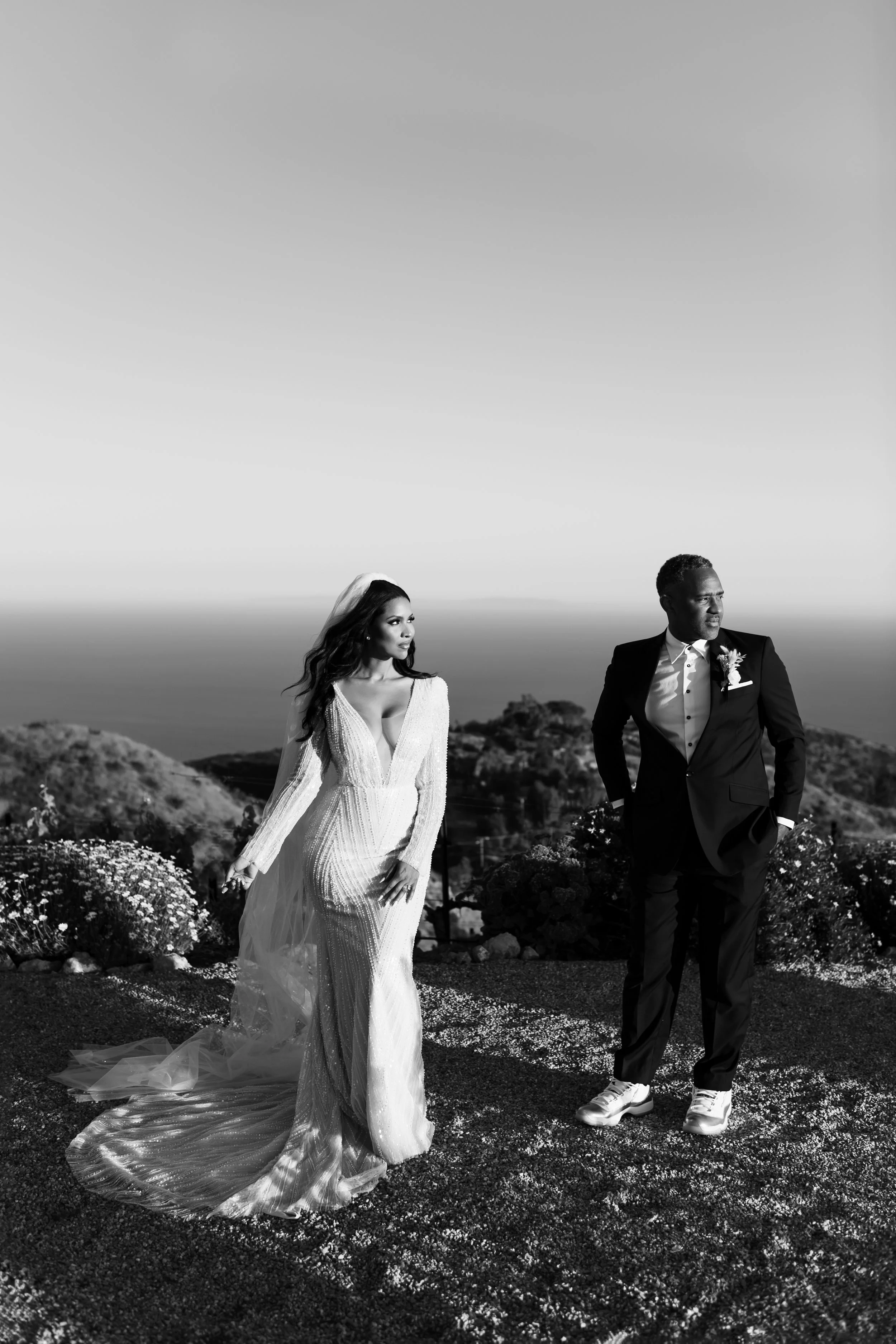 A bride and groom stand outdoors on a rocky, flowered landscape with a distant horizon and sky in the background. The bride wears a long, elegant gown with a veil, and the groom wears a dark tuxedo with a boutonniere and white sneakers. They appear t