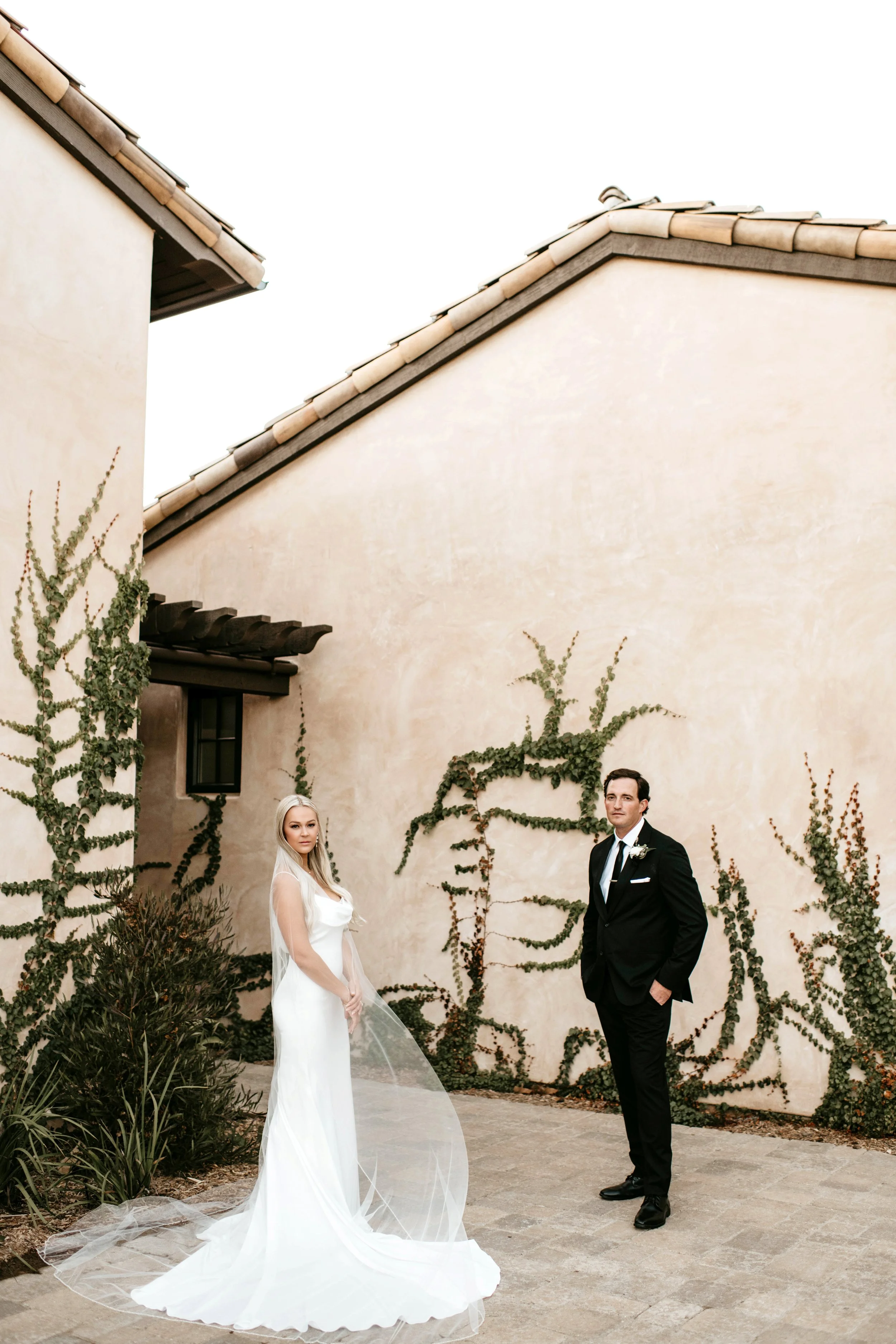 A bride and groom standing outdoors against a beige wall with creeping vines, the bride in a white wedding dress with a veil and the groom in a black tuxedo with a boutonniere.