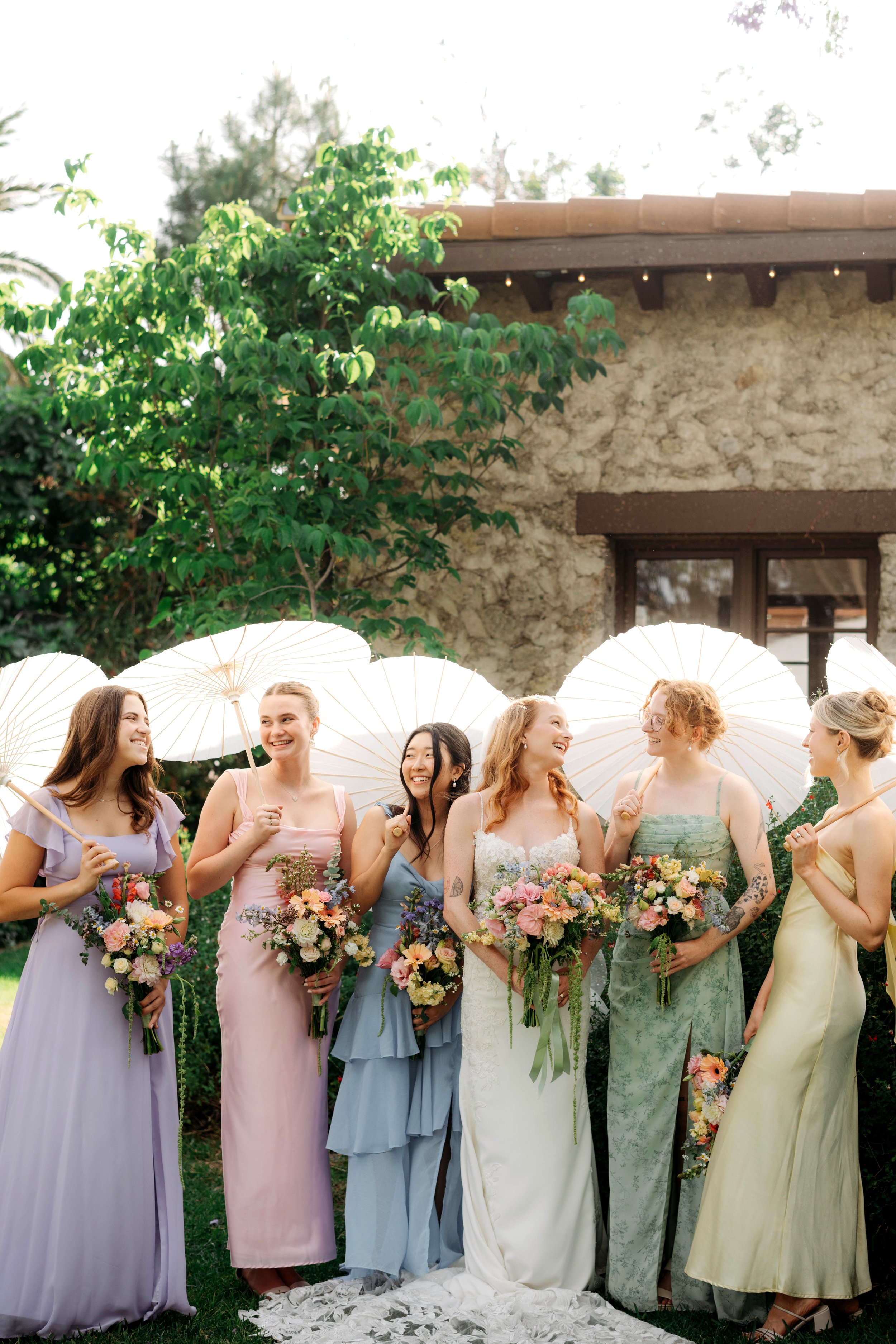 A group of women dressed in pastel-colored dresses holding bouquets of flowers and white umbrellas, smiling and standing in a garden in front of a stone building with a wooden roof overhang and window.