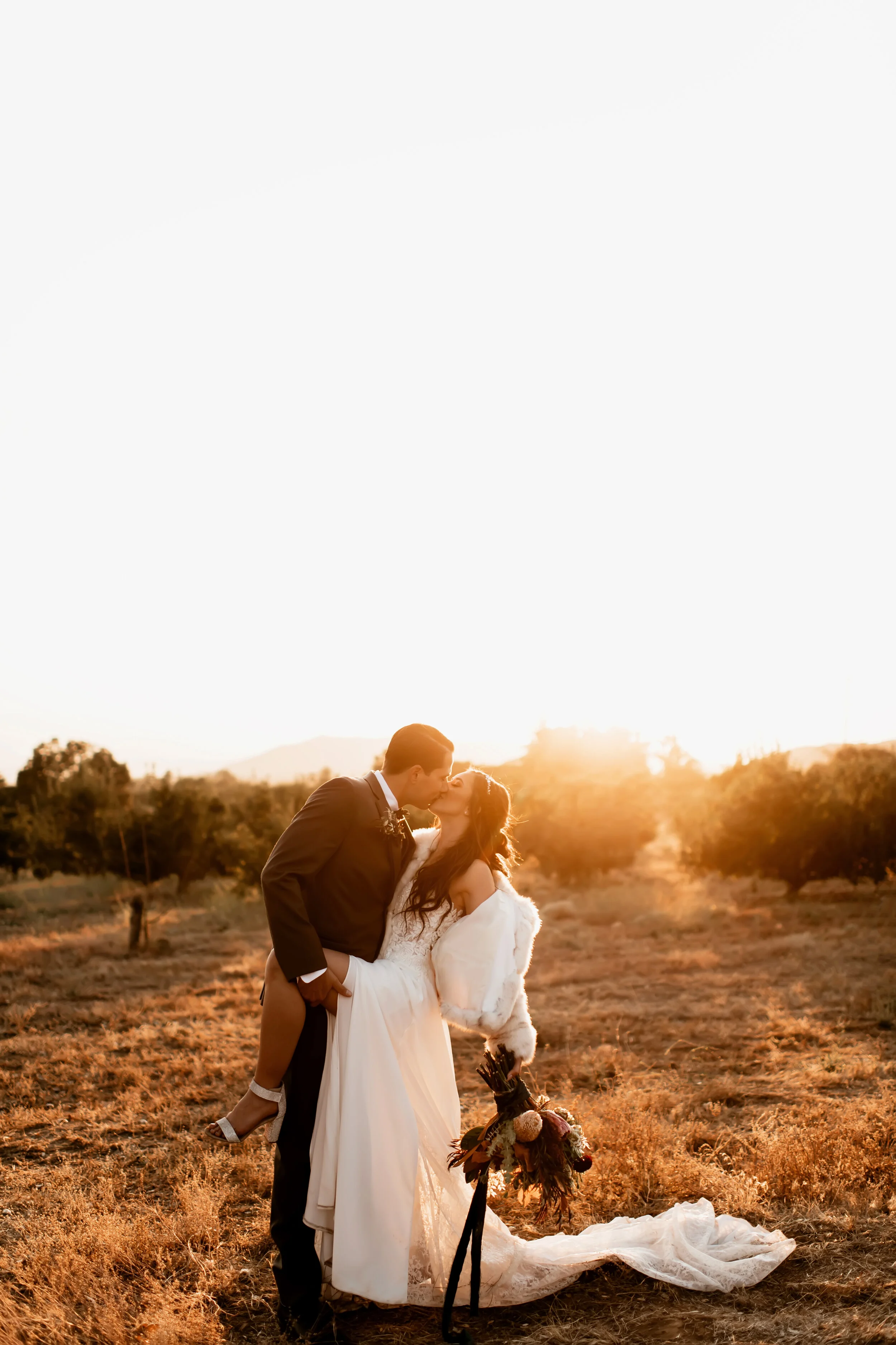 A wedding couple kissing at sunset in an open field. The groom is holding the bride, who is wearing a white dress. The bride holds a bouquet of flowers. The setting sun creates a warm glow over the scene with a few trees and hills in the background.