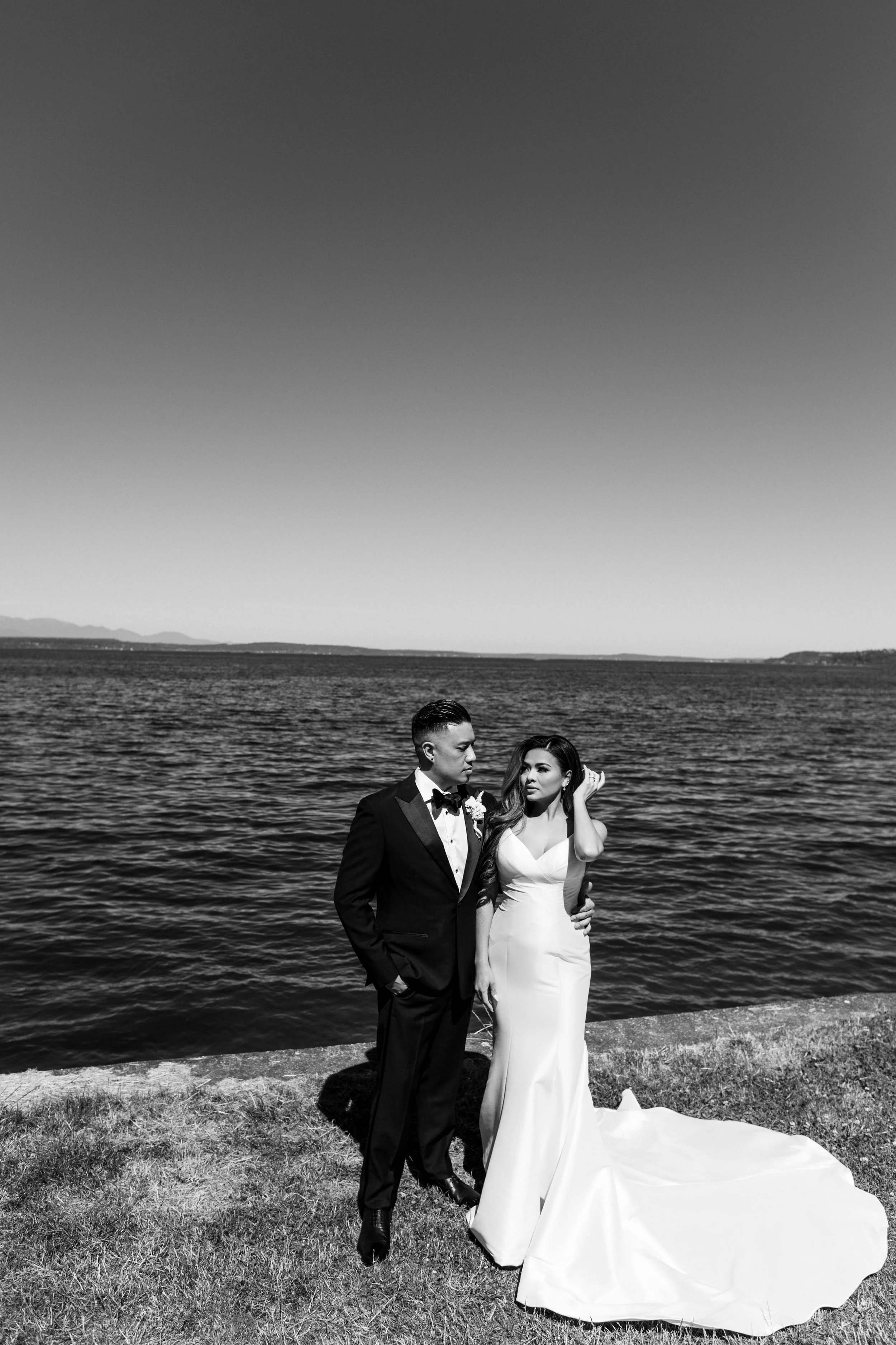 A black and white photograph of a bride and groom standing near a body of water with mountains in the background. The groom is dressed in a tuxedo and the bride in a strapless wedding gown, with her hand touching her hair.