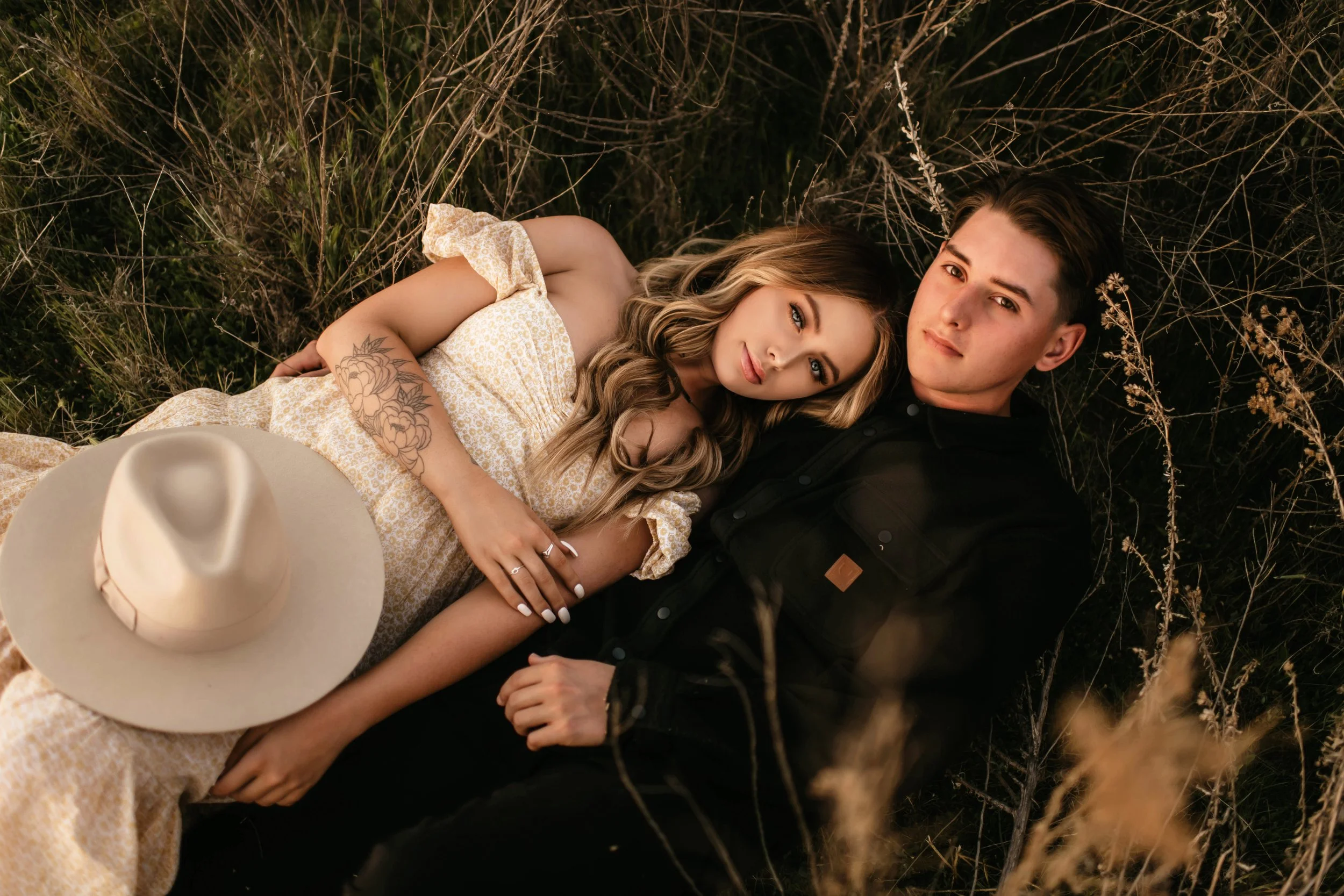 A young woman and young man lying on the ground in a field of dry grass, with the woman resting her head on the man's shoulder, both looking at the camera. The woman is wearing a yellow patterned dress with puffed sleeves, and the man is dressed in a