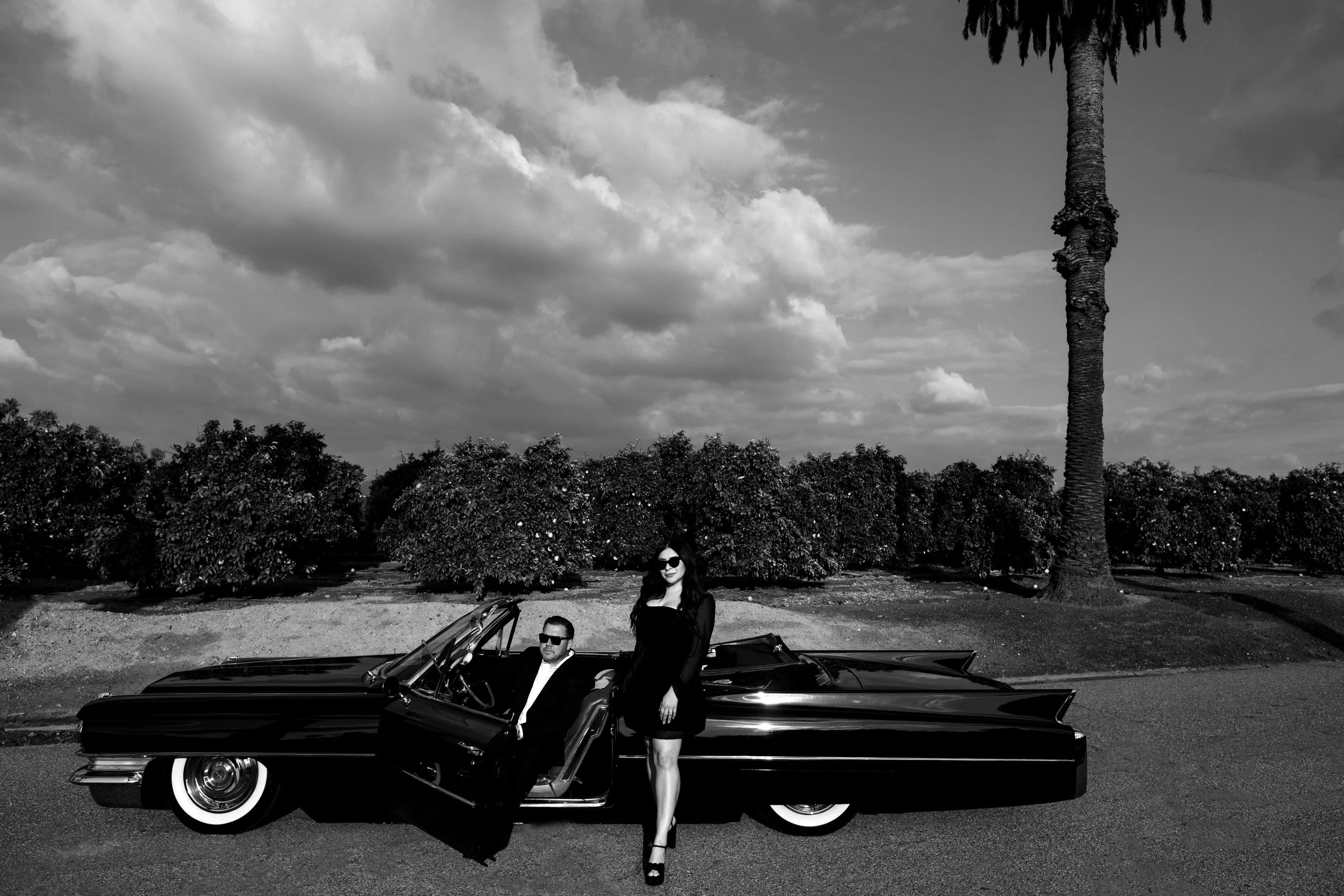 A black and white photo of a man and woman with a vintage convertible car in a park with trees and cloudy sky in the background. The woman is standing outside the car wearing a black dress and high heels, and the man is sitting inside the car wearing