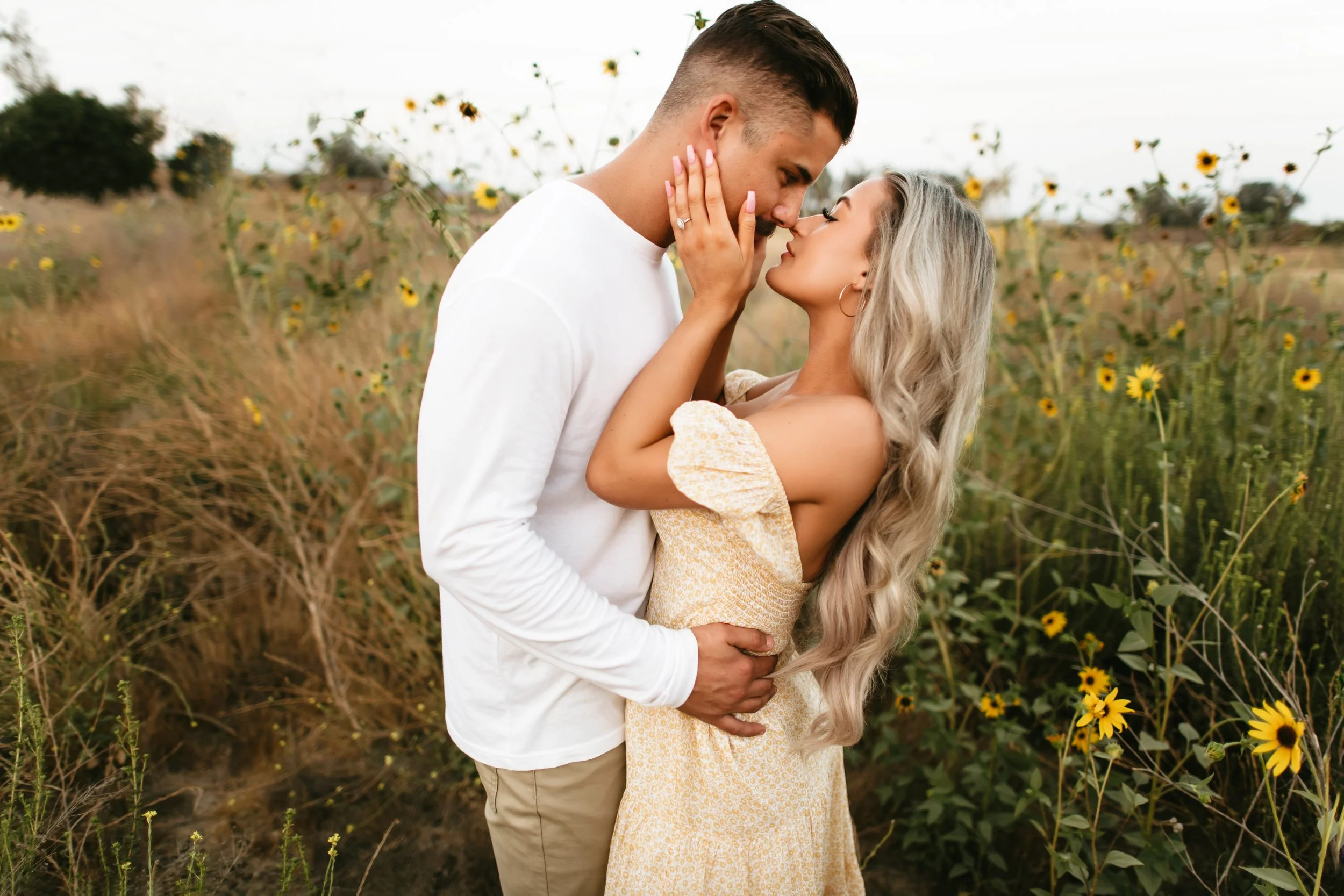 A couple embraces in a sunflower field, about to kiss, with the woman touching the man's face and smiling, both appearing happy.