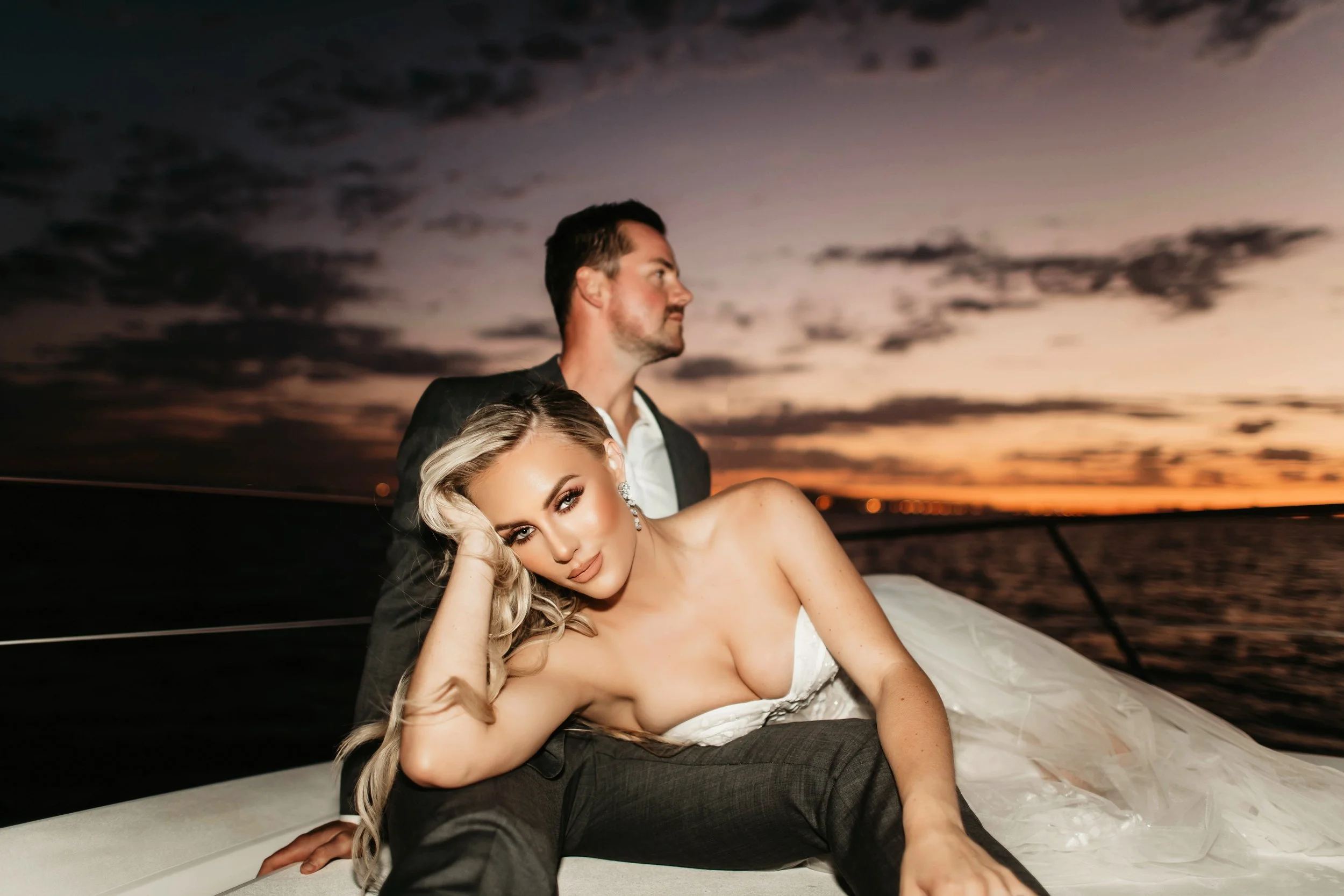 A man and a woman in wedding attire relax on a boat during sunset, with boats on the water and a colorful sky in the background.