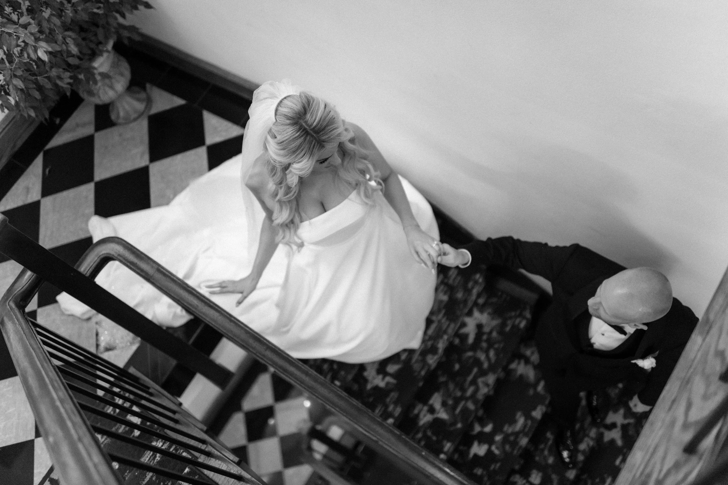 Black and white photo of a woman in a wedding dress with long wavy hair, and a man in formal attire, sitting on a bed in a room with checkered floor tiles.