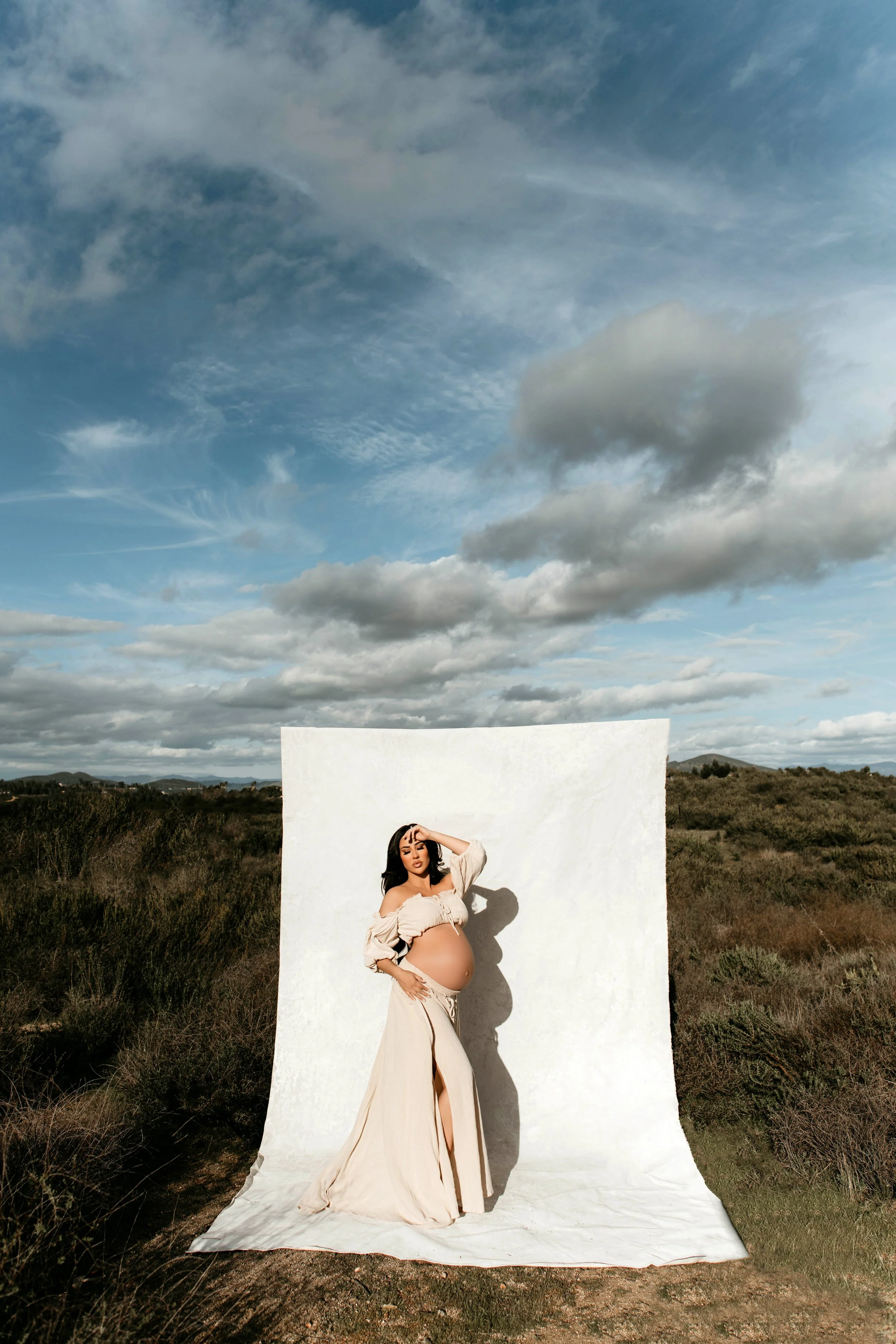 A pregnant woman standing outdoors in front of a white backdrop, holding her belly, with cloudy sky and open landscape behind her.