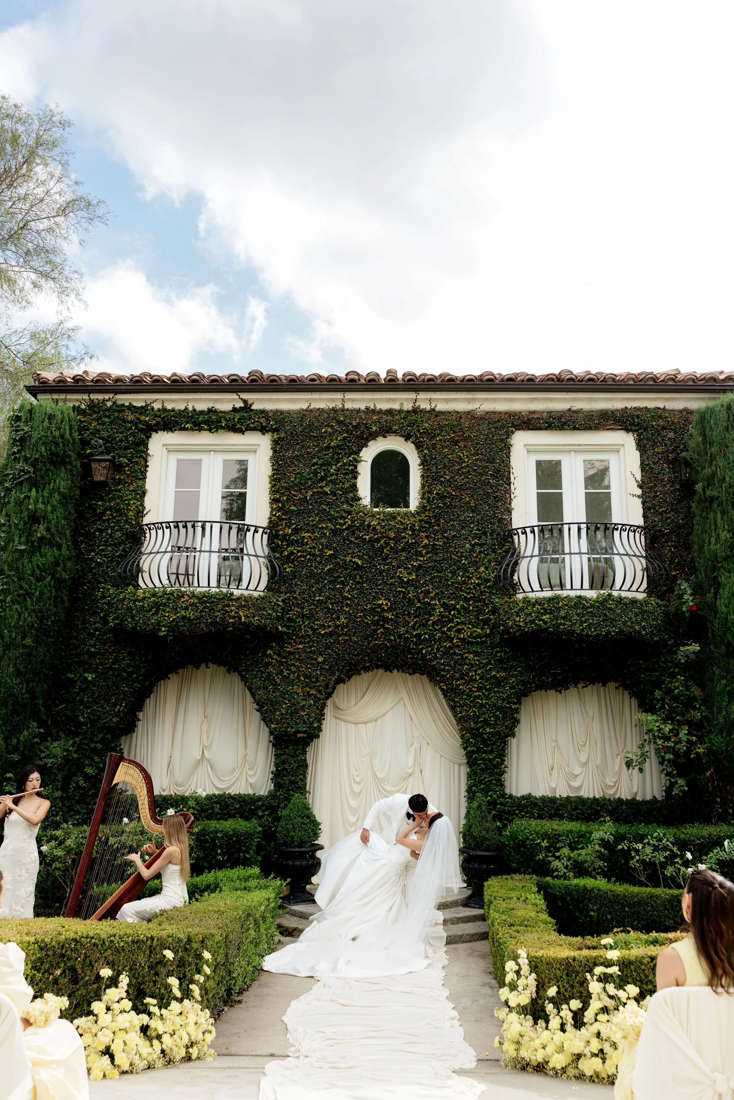 A wedding ceremony taking place outdoors in front of a green ivy-covered building with two balconies, where a groom is kissing the bride. Musicians are playing, and guests are seated along a white floral aisle.