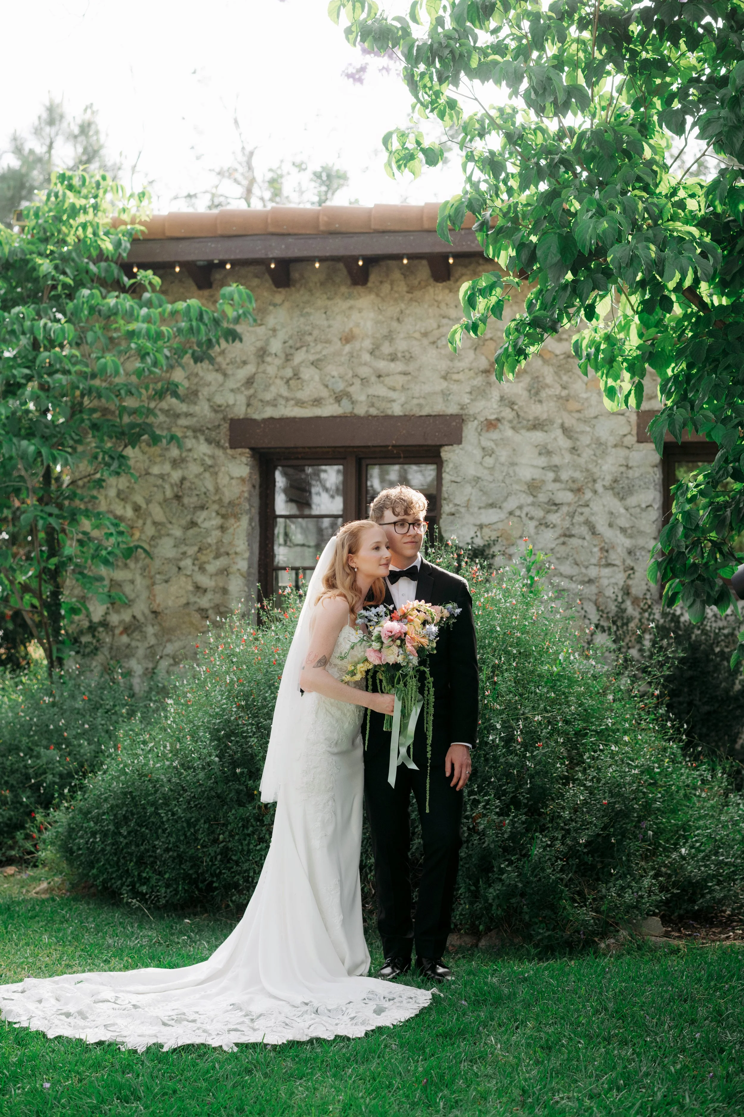A newlywed couple, a woman in a white wedding gown holding a bouquet and a man in a black tuxedo, stands together in a lush garden with greenery and flowers, in front of a stone building with windows and a tiled roof.