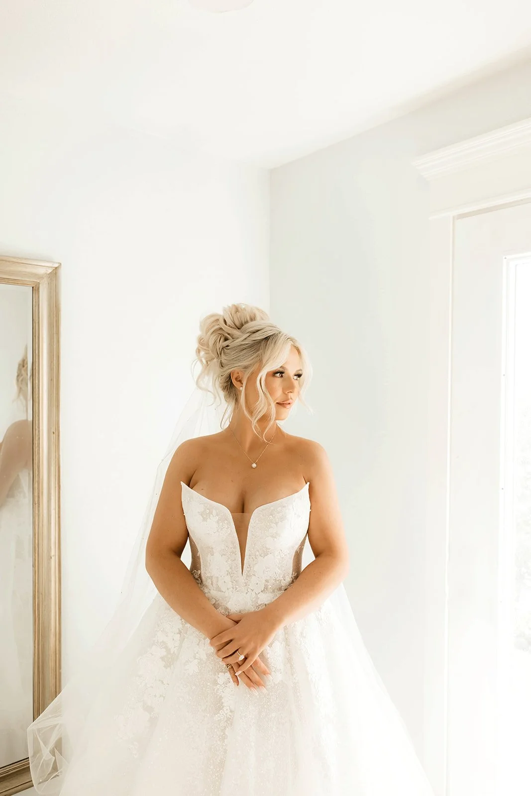 Bride in a white wedding gown standing near a mirror in a bright room