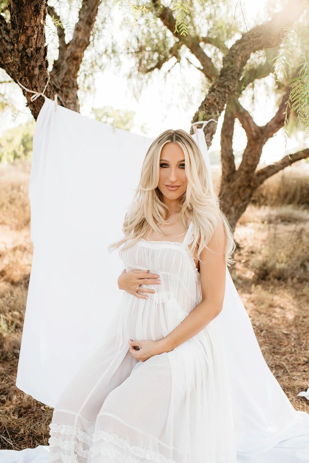 A woman in a white dress sitting outdoors next to a tree with a white sheet hanging on a line behind her, illuminated by warm sunlight.