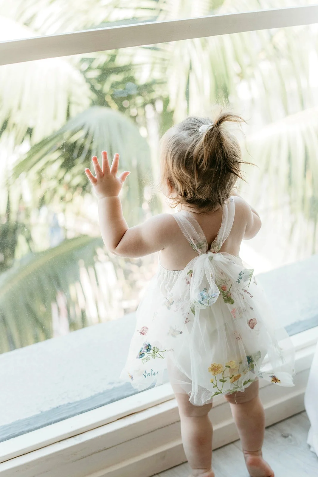 A young girl with short curly hair, wearing a white dress with a floral pattern and bow, stands by a large window with her hand touching the glass, looking outside at green palm trees.