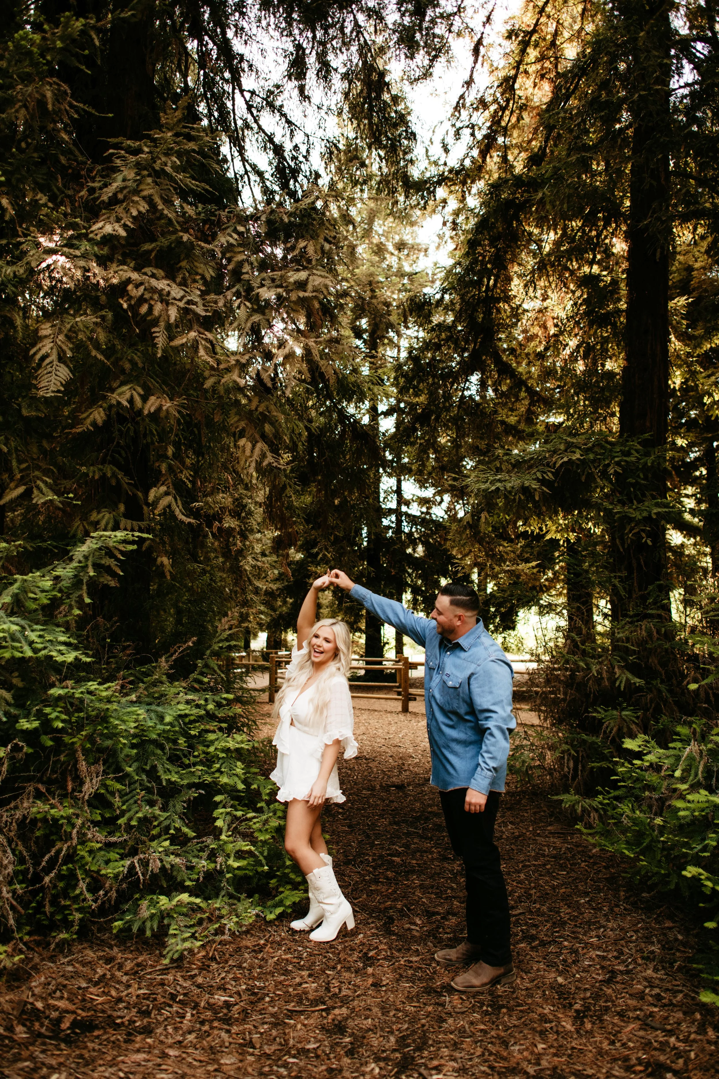 A man and woman dancing in a forest, holding hands, surrounded by tall trees with green foliage and sunlight filtering through the branches.