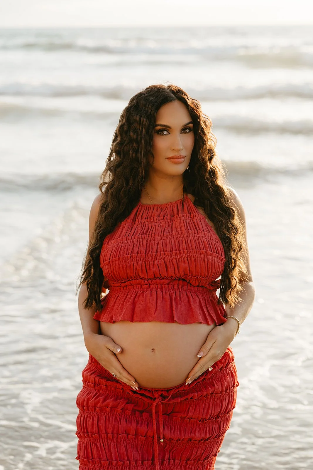 A pregnant woman in a red ruffled top and matching pants stands on the beach with ocean waves in the background, gently holding her belly.