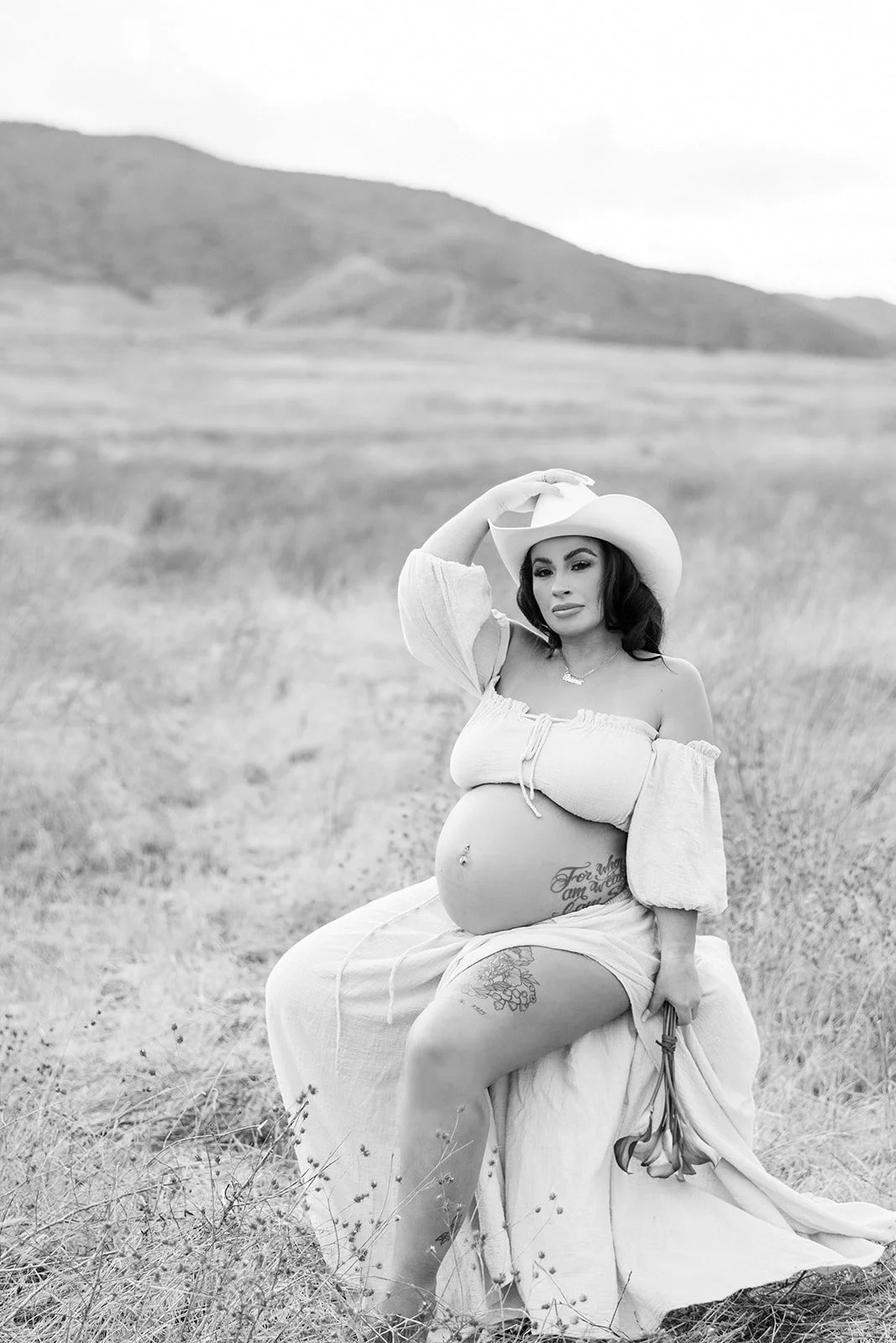 A pregnant woman in a wide-brimmed hat sitting in a field, holding a bouquet of flowers in one hand, with mountains in the background.